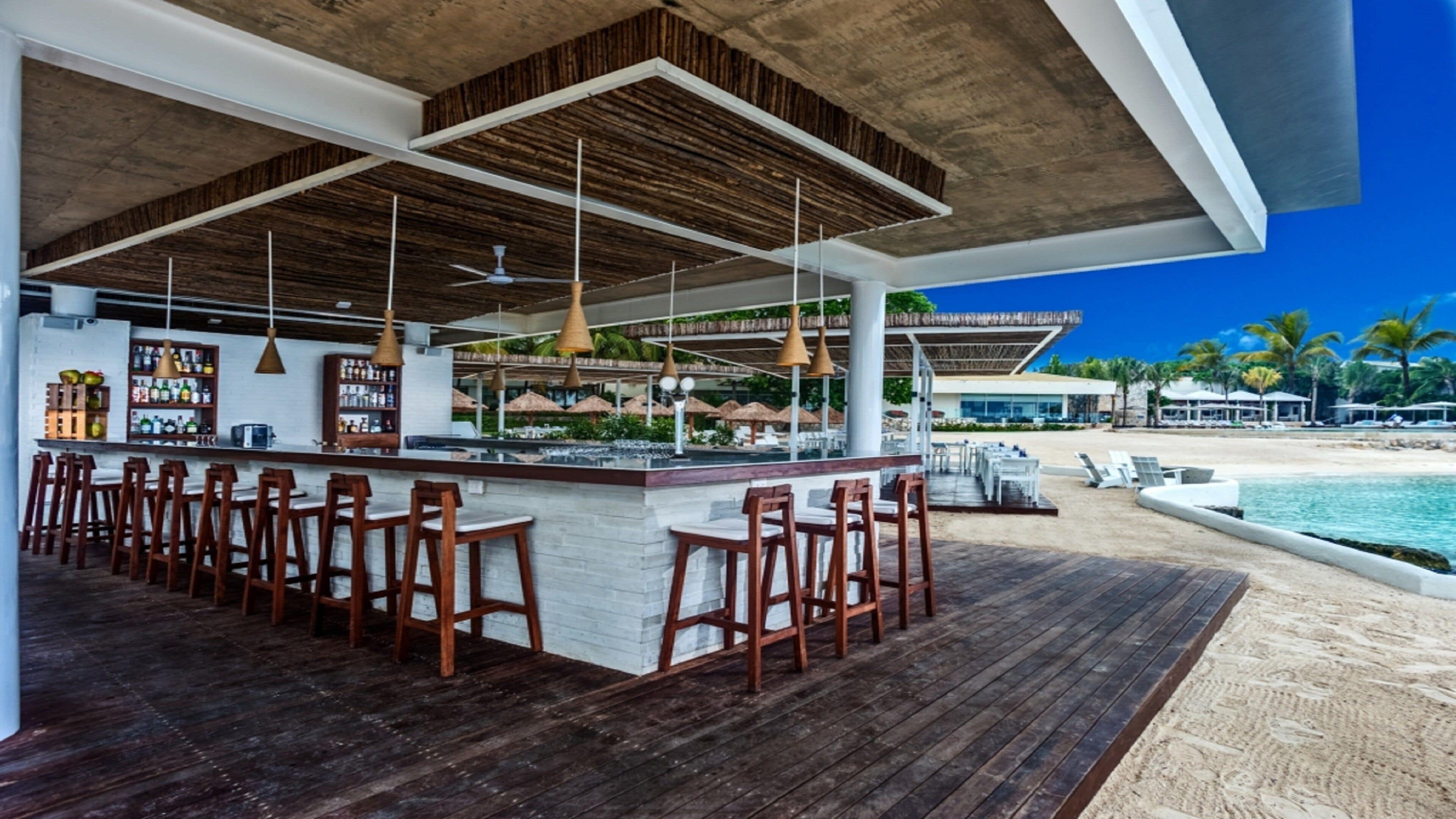 A bar area with wooden chairs and a white counter, overlooking a beach with umbrellas and lounge chairs.