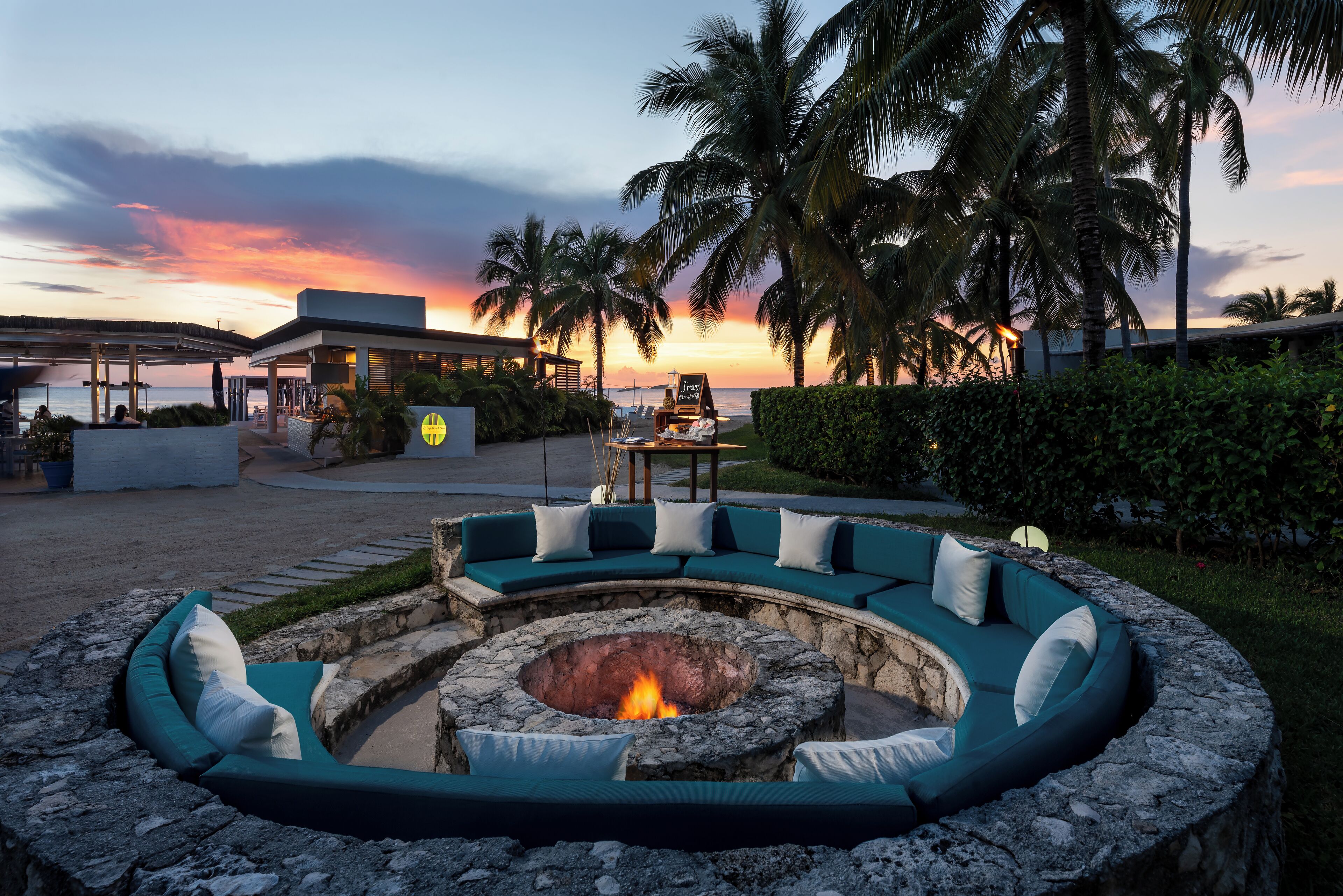 A circular outdoor seating area with a fire pit, surrounded by a stone wall and a curved green sofa with white cushions.