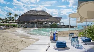 Een eetgedeelte aan het strand met een tafel voor twee, met uitzicht op de oceaan en een gebouw met rieten dak.