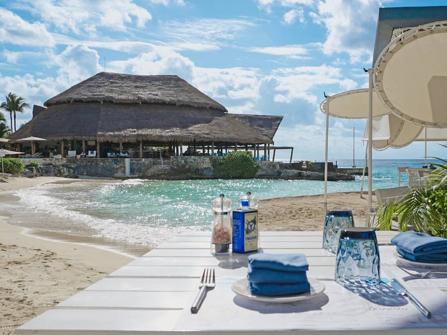 Een eetgedeelte aan het strand met een tafel voor twee, met uitzicht op de oceaan en een gebouw met rieten dak.