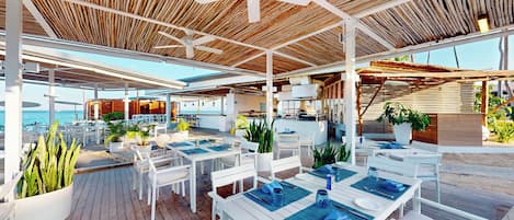 An outdoor dining area with white tables and chairs, a wooden ceiling, and a view of the sea.