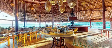 A spacious indoor area with a high ceiling, wooden beams, and a thatched roof. There are tables and chairs arranged for dining, and a large window offering a view of the ocean.