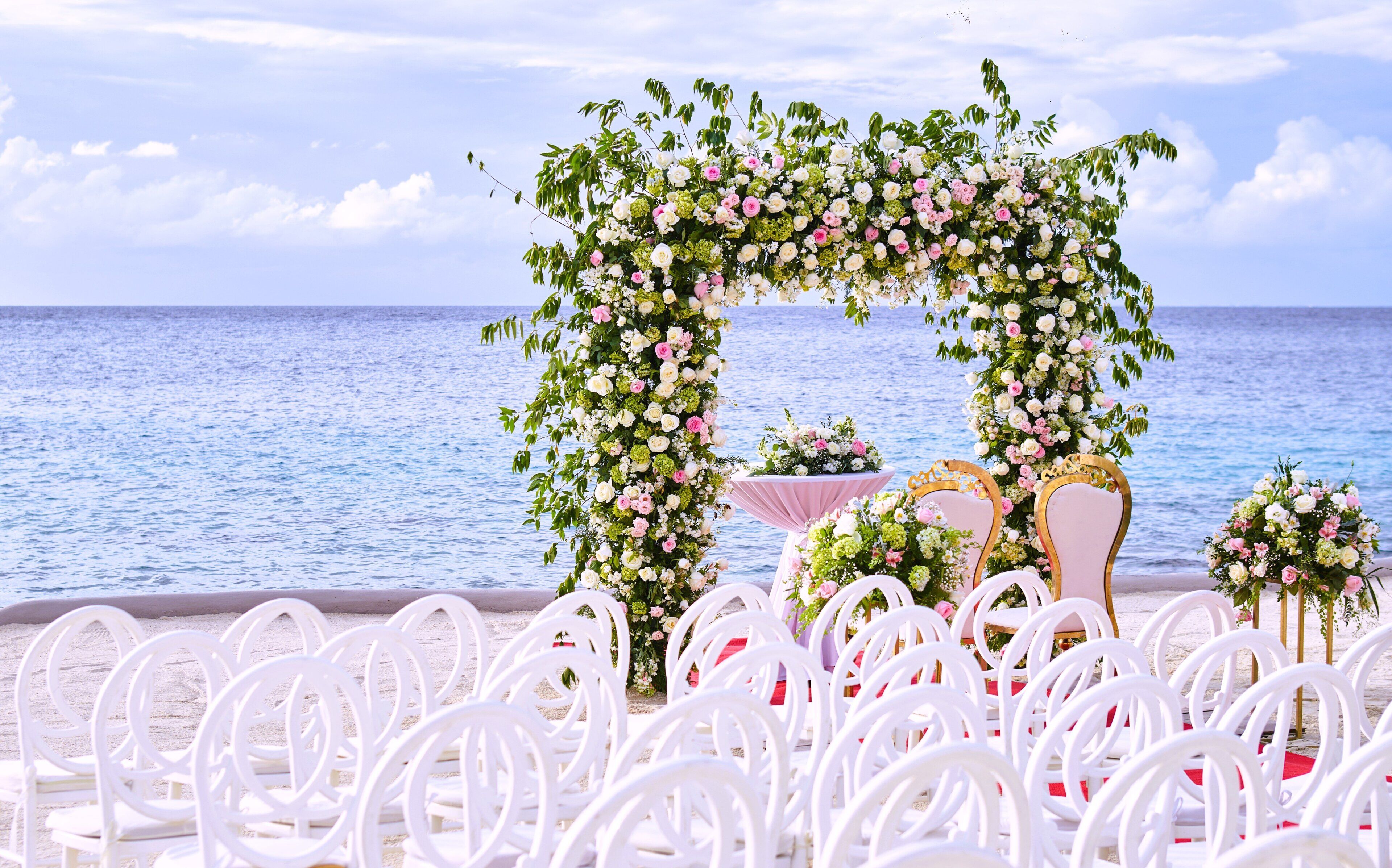A beachside wedding setup with white chairs and a floral arch.