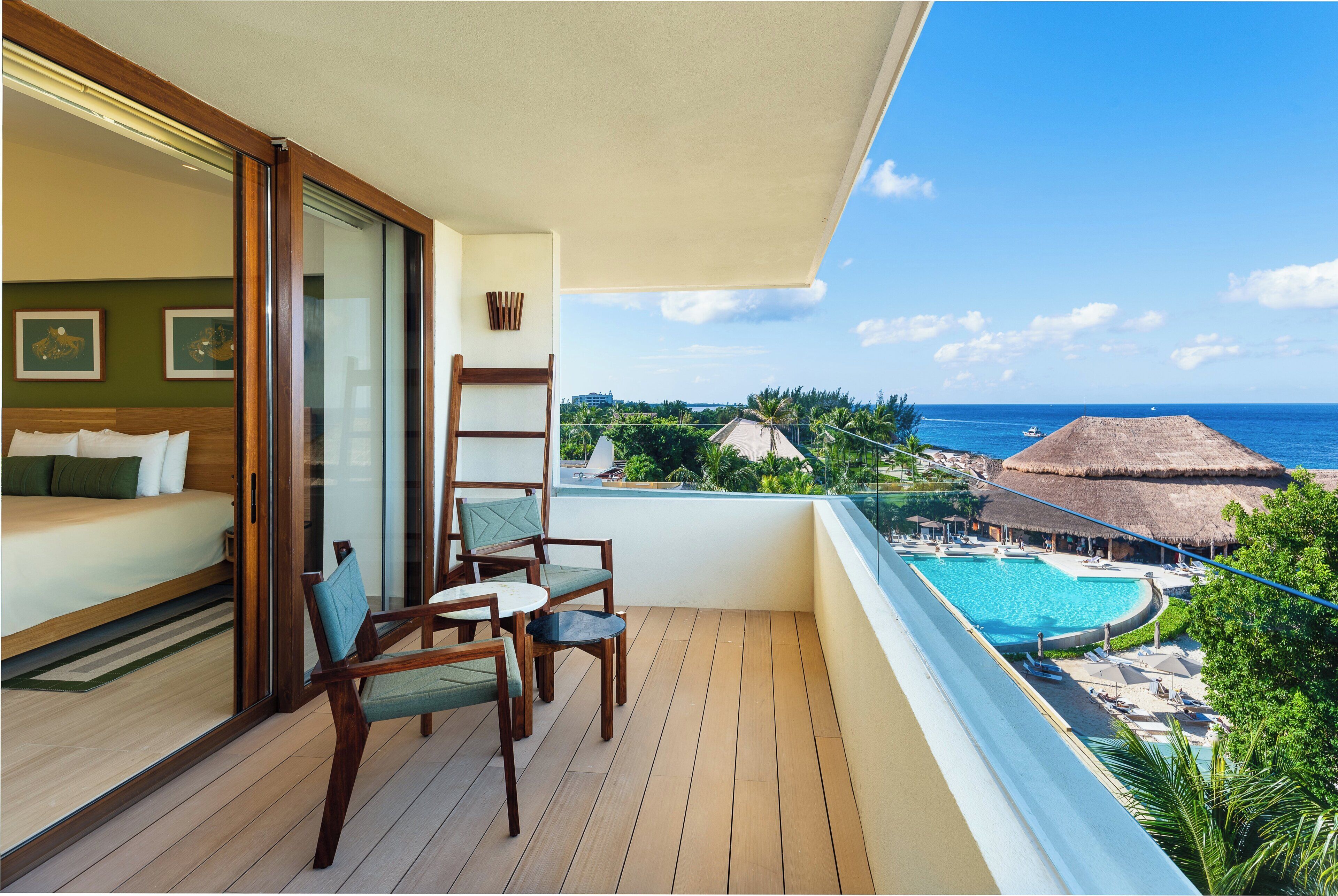 Uma varanda com piso de madeira, uma cama com almofadas verdes, uma cadeira e vista para uma piscina e construções com telhado de palha.