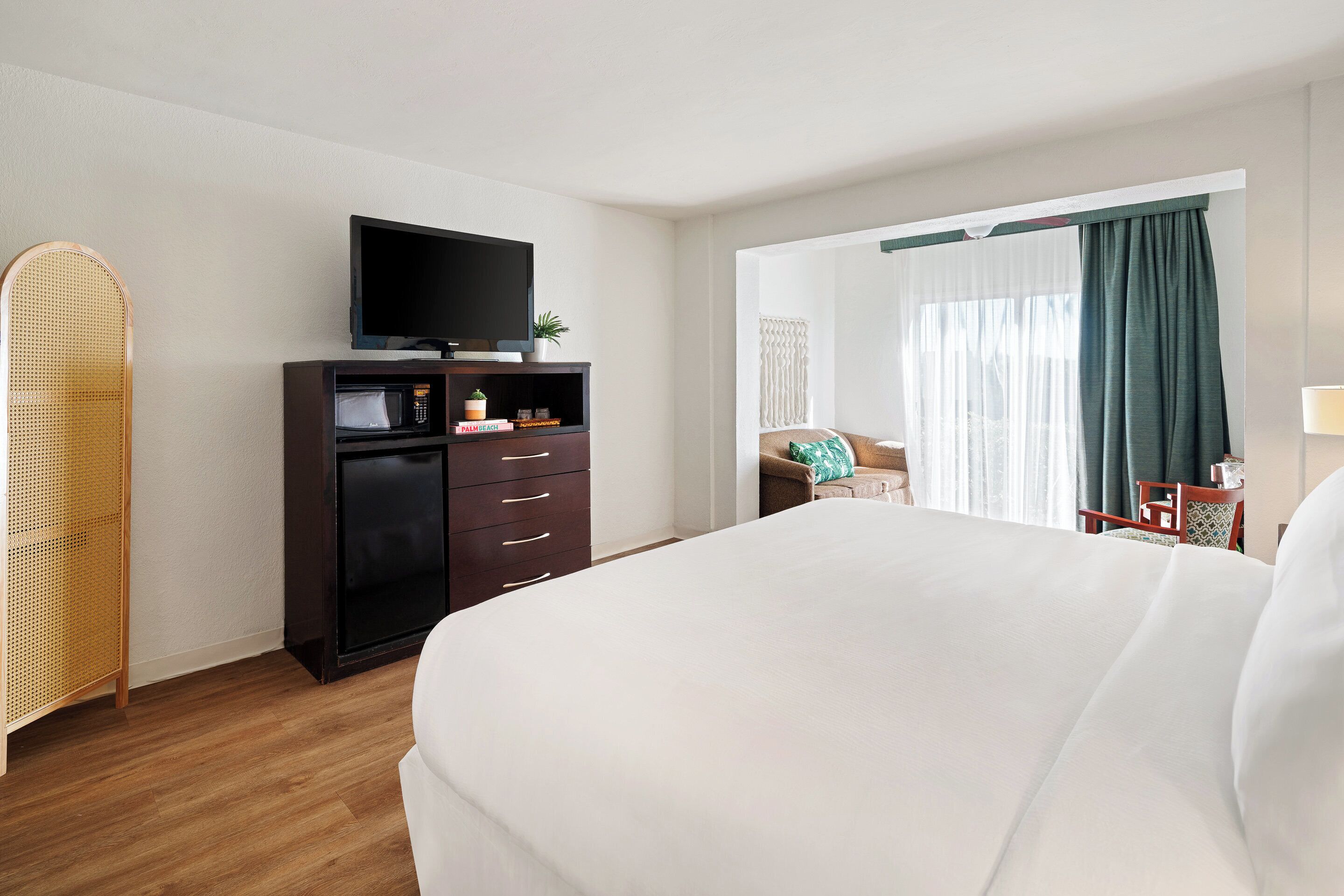 Quarto de hotel com uma cama grande, televisão de tela plana em um suporte, minibar, cadeira de vime e vista para o exterior através de uma janela com cortinas.