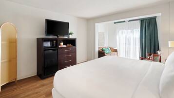 Quarto de hotel com uma cama grande, televisão de tela plana em um suporte, minibar, cadeira de vime e vista para o exterior através de uma janela com cortinas.