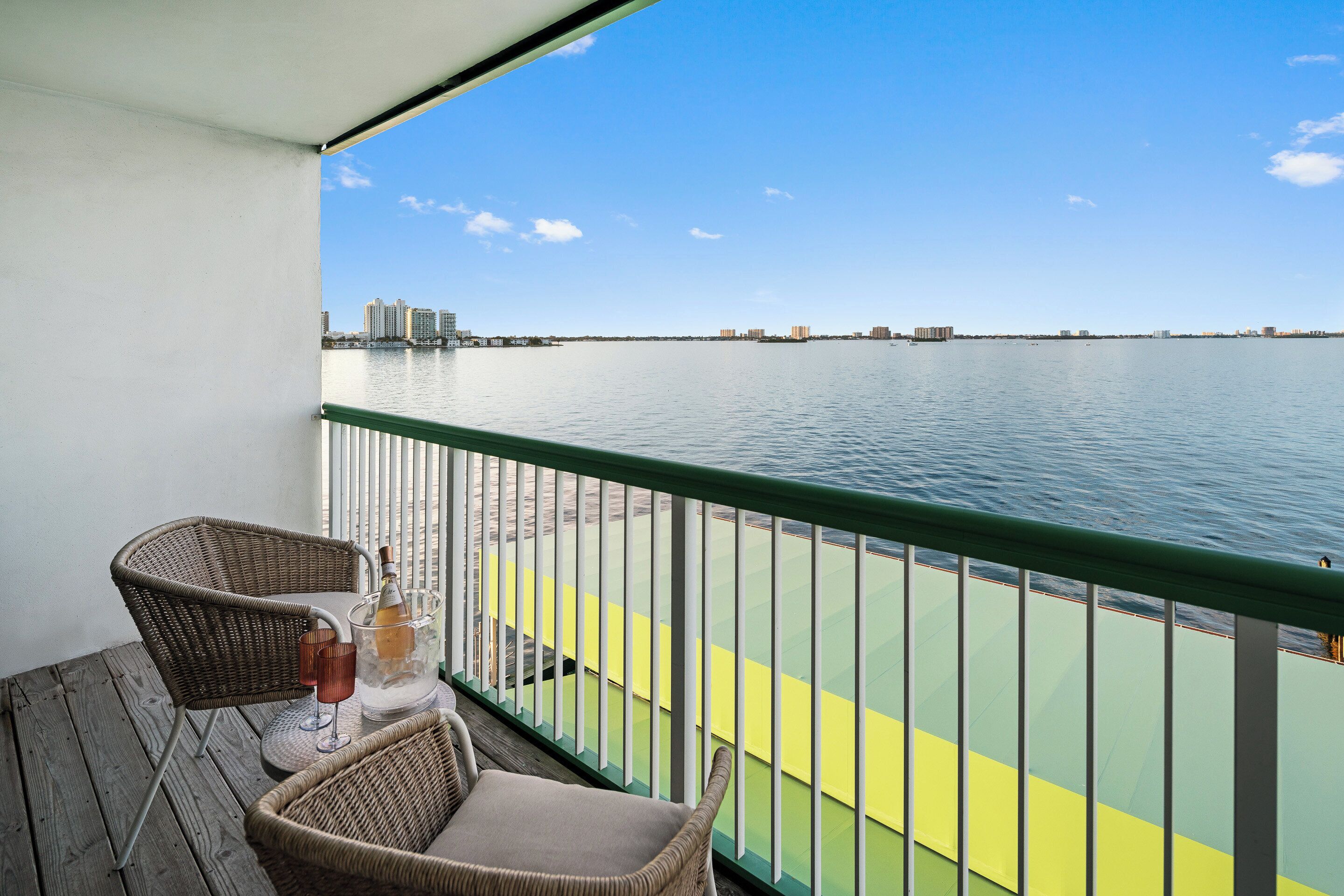 A balcony with wicker chairs, a table with a bottle and glass, and a view of the water and distant buildings.