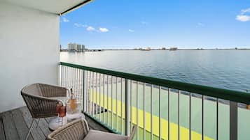 A balcony with wicker chairs, a table with a bottle and glass, and a view of the water and distant buildings.