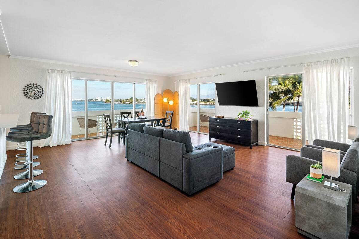 A modern living room with a large gray sectional sofa, a dining area with a table and chairs, a flat-screen TV mounted on the wall, and a view of the ocean through large windows.