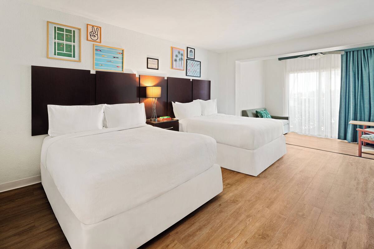 A hotel room with two beds, a nightstand with a lamp, and a view of the outdoors through a window with curtains.