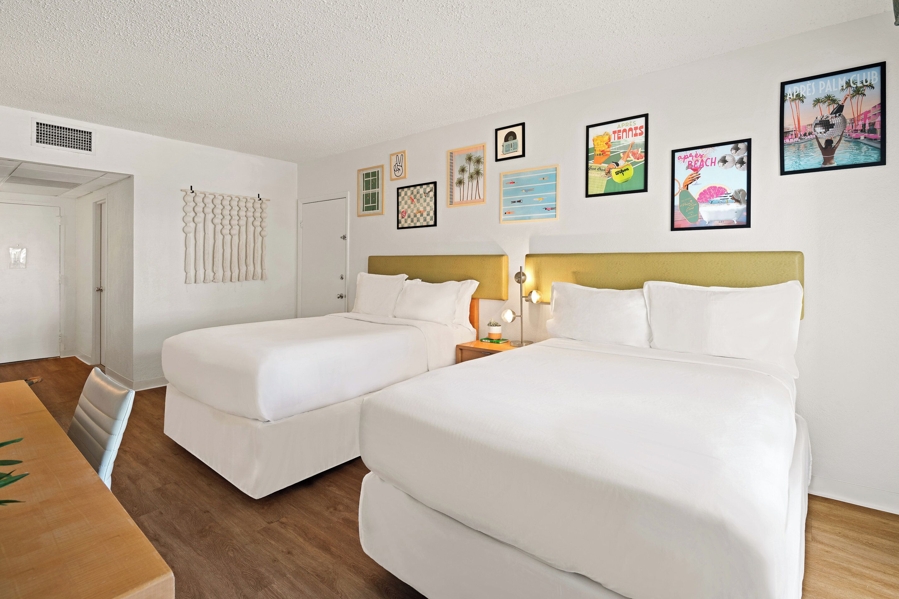 Une chambre d’h ôtel avec deux lits simples, une table en bois et un mur décoré de divers tableaux encadrés.