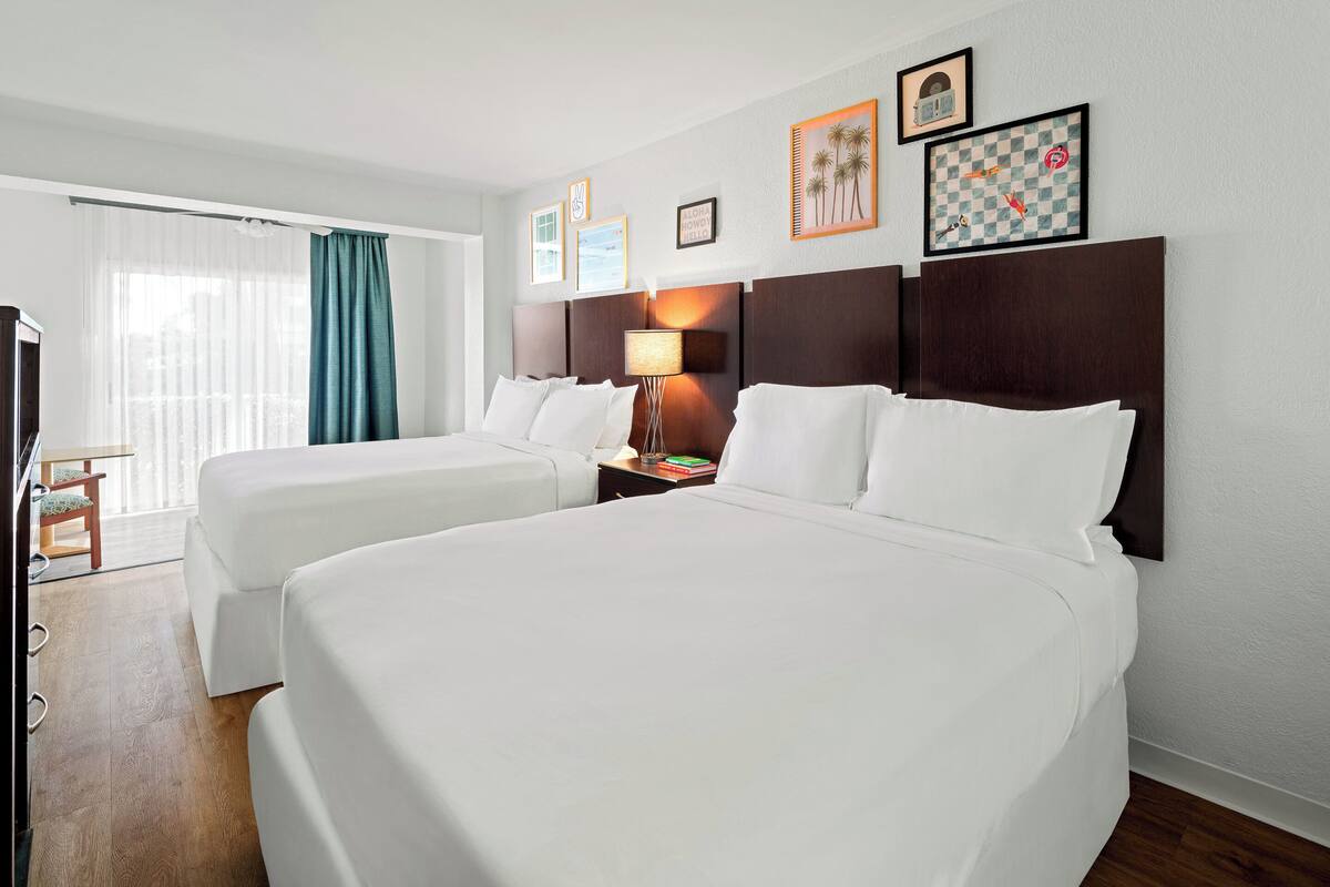 A hotel room with two beds, a desk, a chair, a lamp, and framed pictures on the wall.