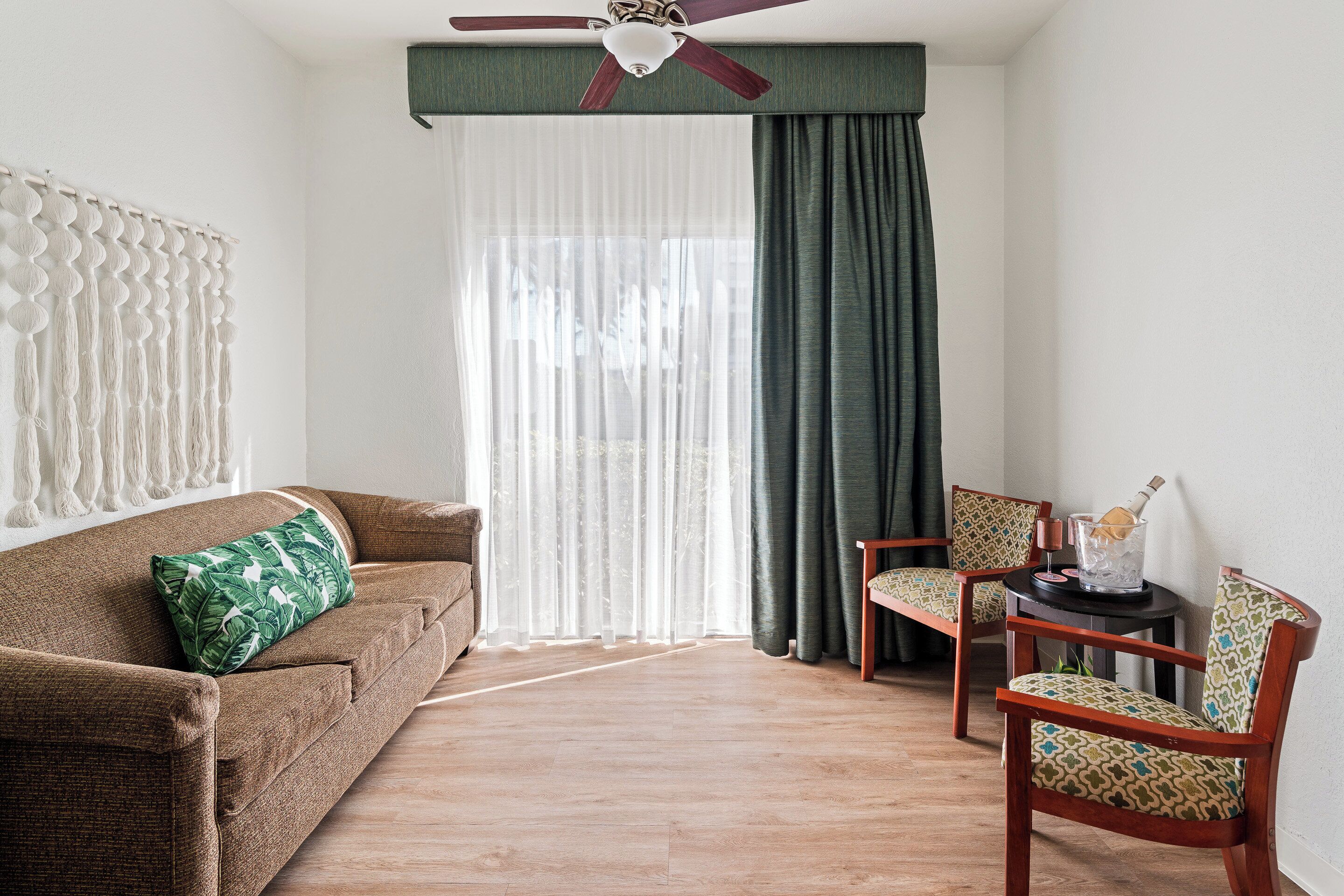 Un salón con un sofá marrón, un cojín verde estampado, dos sillas de madera, un ventilador de techo y una ventana con cortinas traslúcidas y un drapo oscuro.