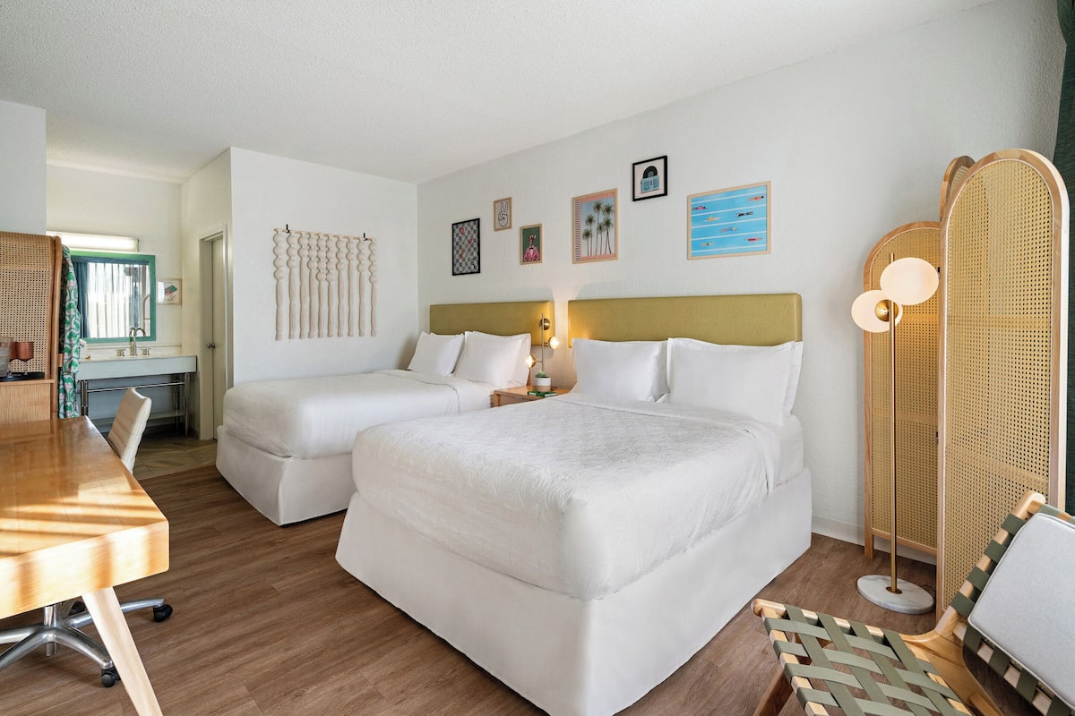 A hotel room with two beds, a desk, a chair, and a large window.