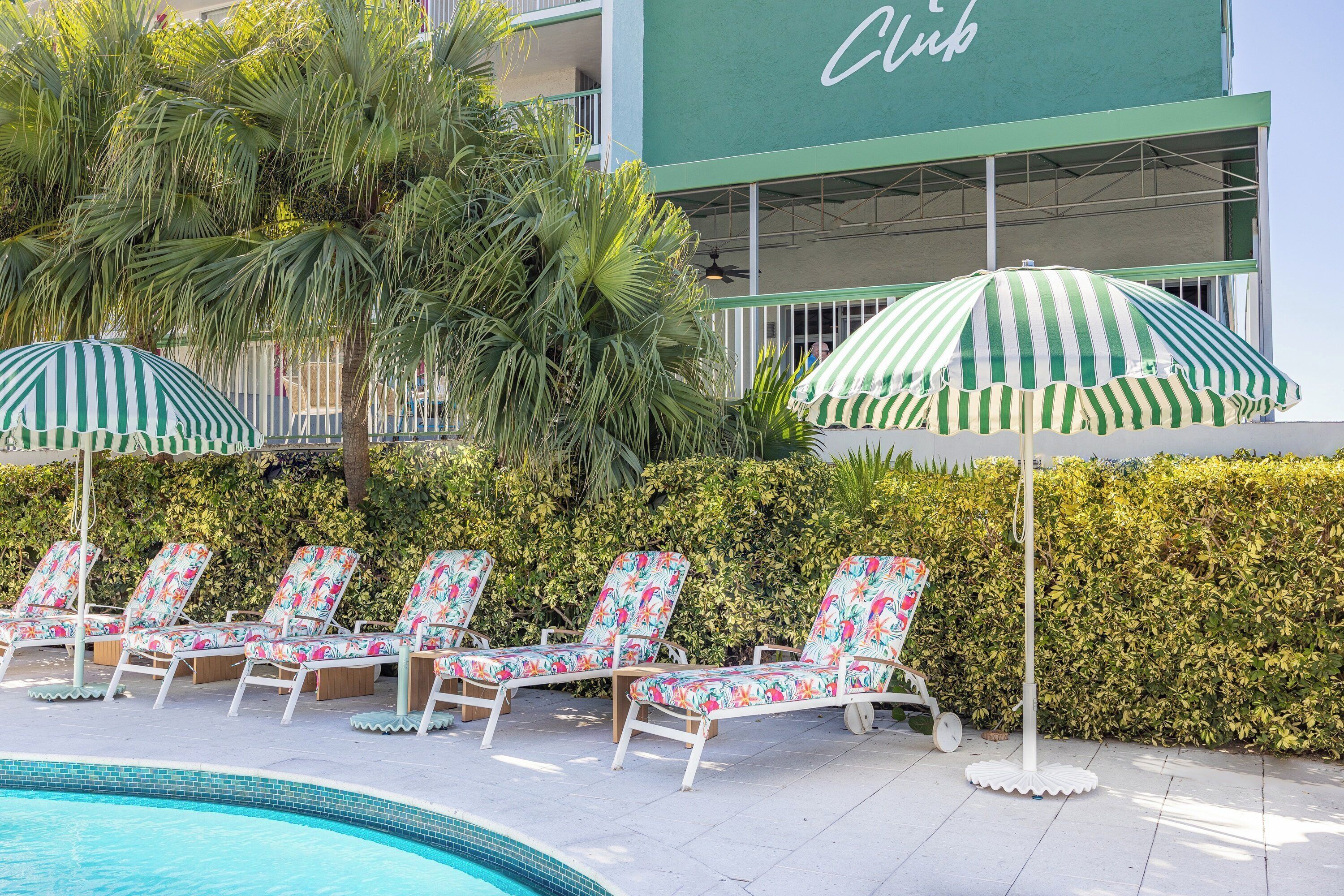 A poolside area with lounge chairs and striped umbrellas, a building with a 'Club' sign, and a well-maintained hedge in the background.