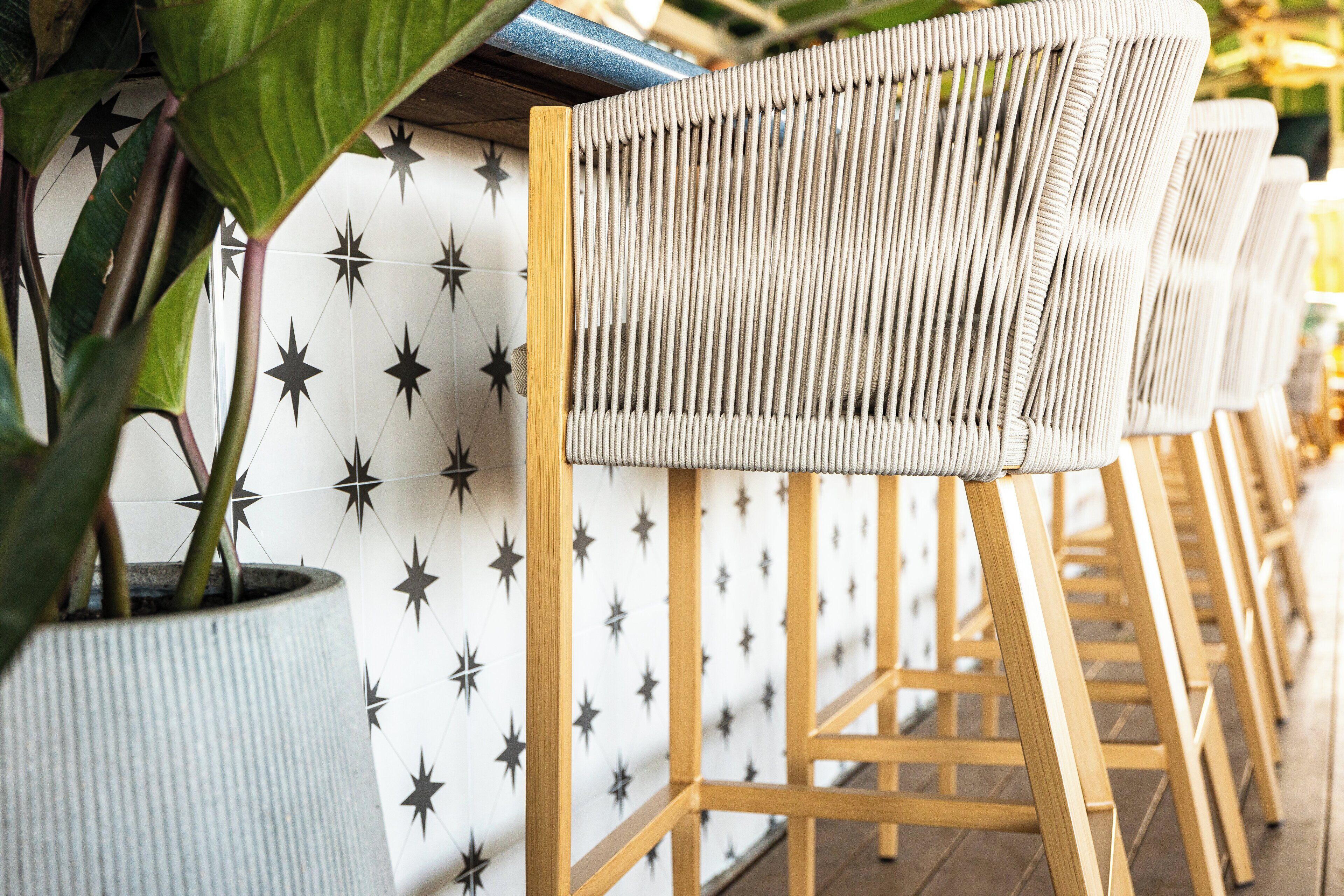 Une rangée de chaises en osier, dotées de pieds en bois et d’un coussin blanc, est alignée contre un mur orné d’un motif en étoile.