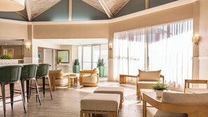 A spacious hotel lobby with wooden flooring, a seating area with ottomans and chairs, a bar with wicker chairs, and large windows with sheer curtains. - Palm Tree Club Miami Hotel (North Bay Village)