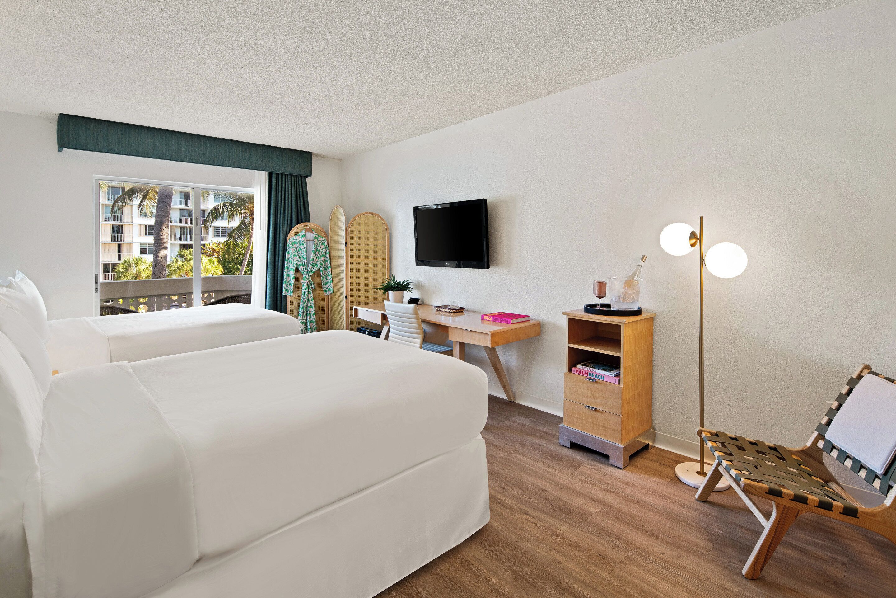 A hotel room with a bed, a desk, a chair, a TV, a bookshelf, and a window with a view of palm trees.