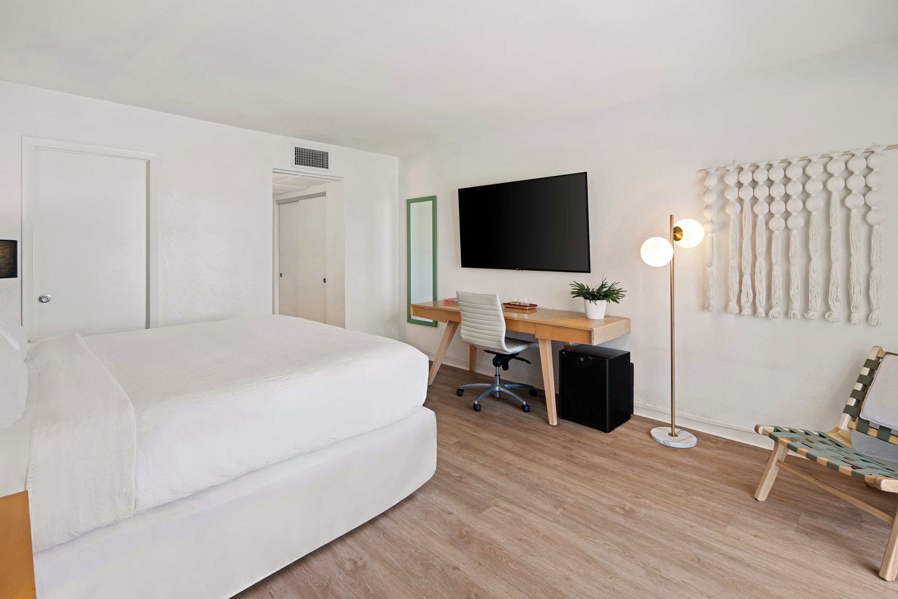 A modern bedroom with a large bed, a desk with a chair, a flat-screen TV, a floor lamp, and a decorative wall hanging.