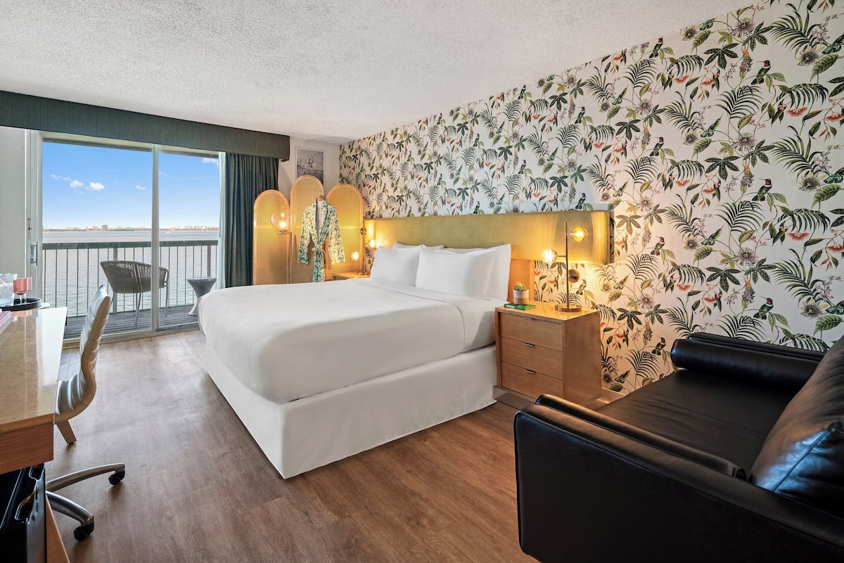 A hotel room with a large bed, a desk, a chair, a sofa, a balcony with a view, and tropical wallpaper.