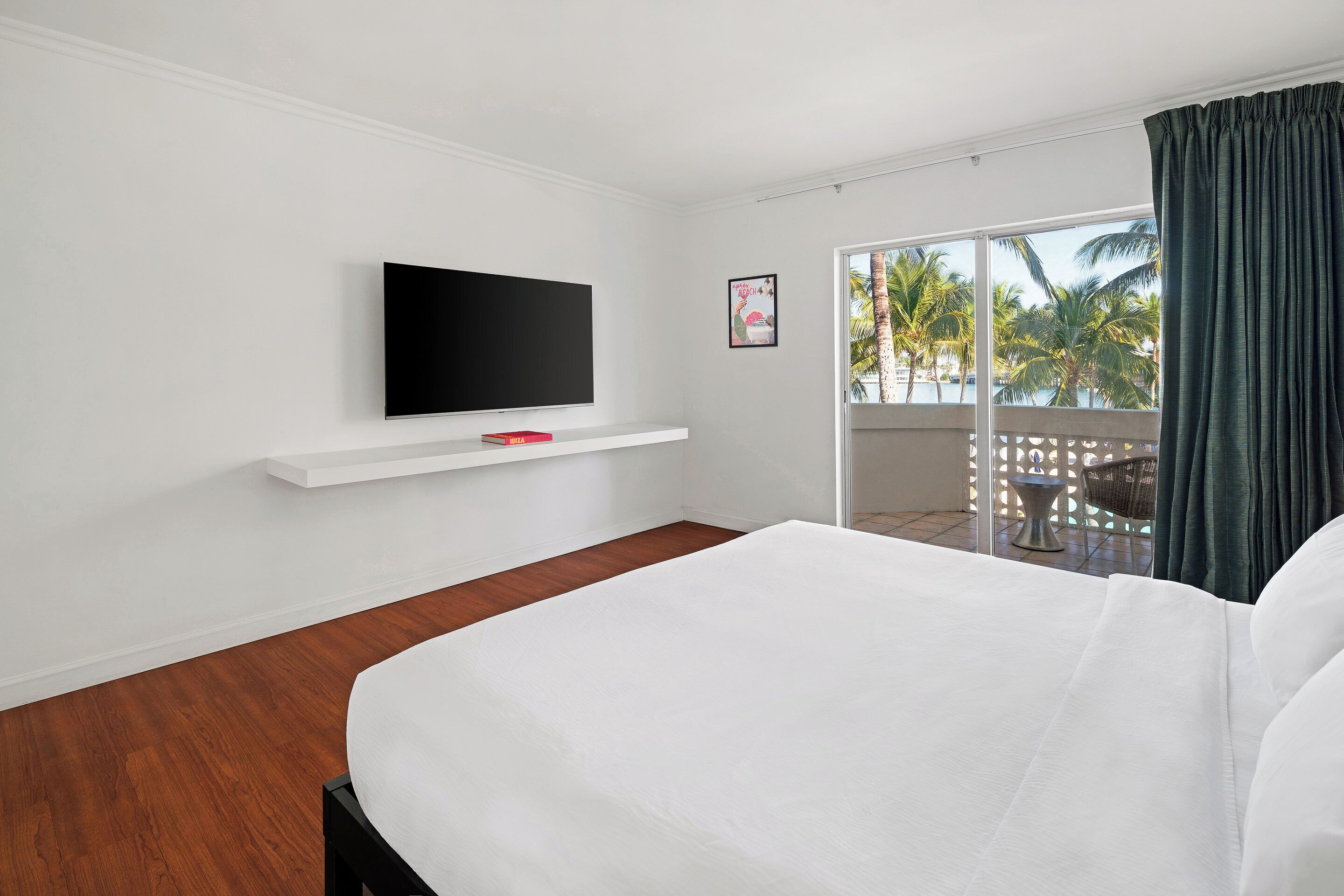 Ein Hotelzimmer mit Bett, einem angebrachten Fernseher, einer kleinen Ablage und einem Balkon mit Blick auf Palmen.