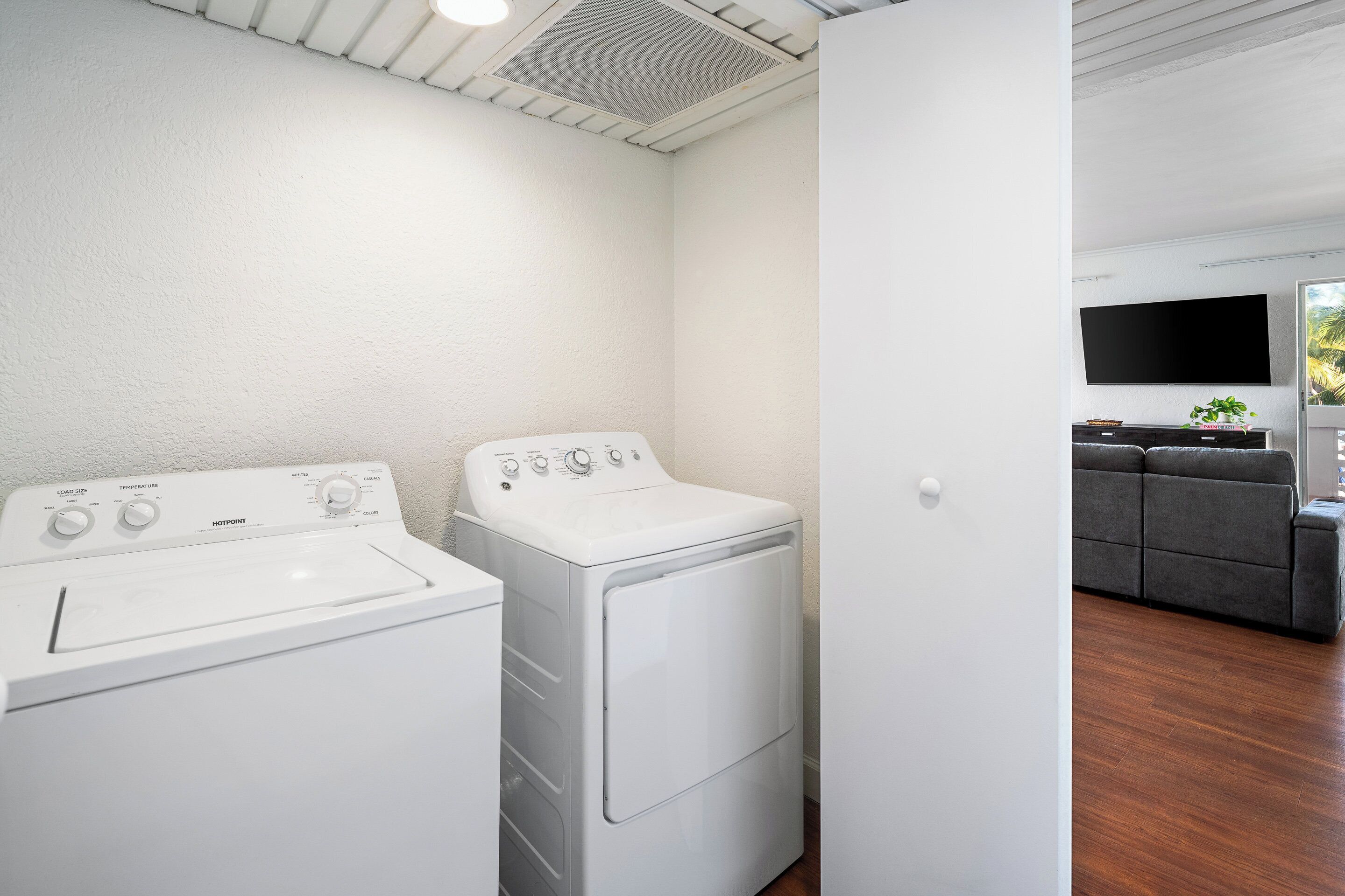 Une buanderie équipée d’un lave-linge et d’un sèche-linge à chargement frontal, d’un canapé gris et d’un téléviseur à écran plat fixé au mur.