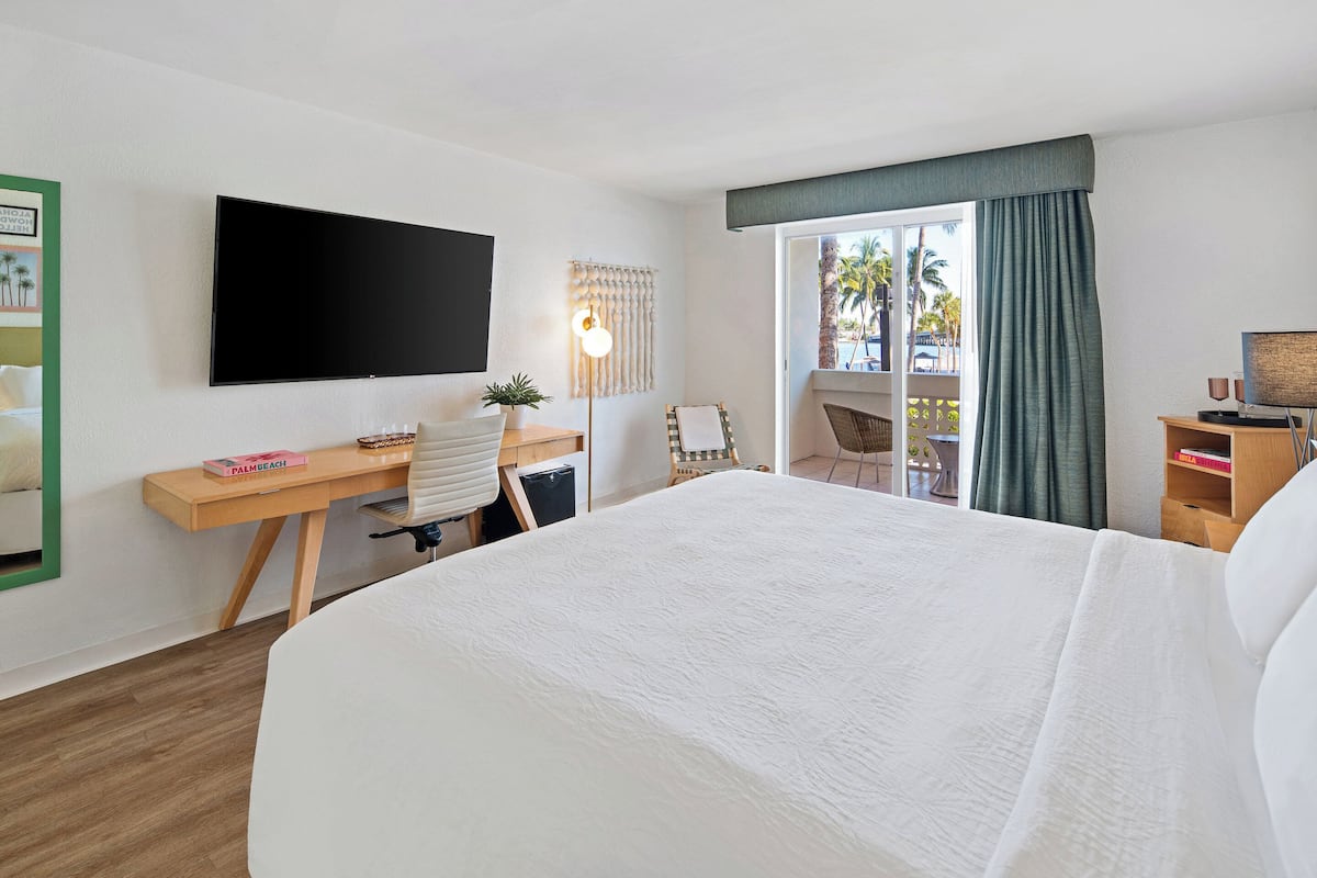 A hotel room with a large bed, a desk with a computer, a flat-screen TV, and a view of palm trees through the window.