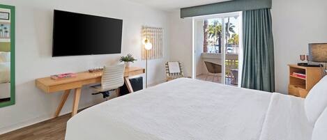 Habitación de hotel con una cama grande, un escritorio con computadora, un televisor de pantalla plana y vista a palmeras a través de la ventana.
