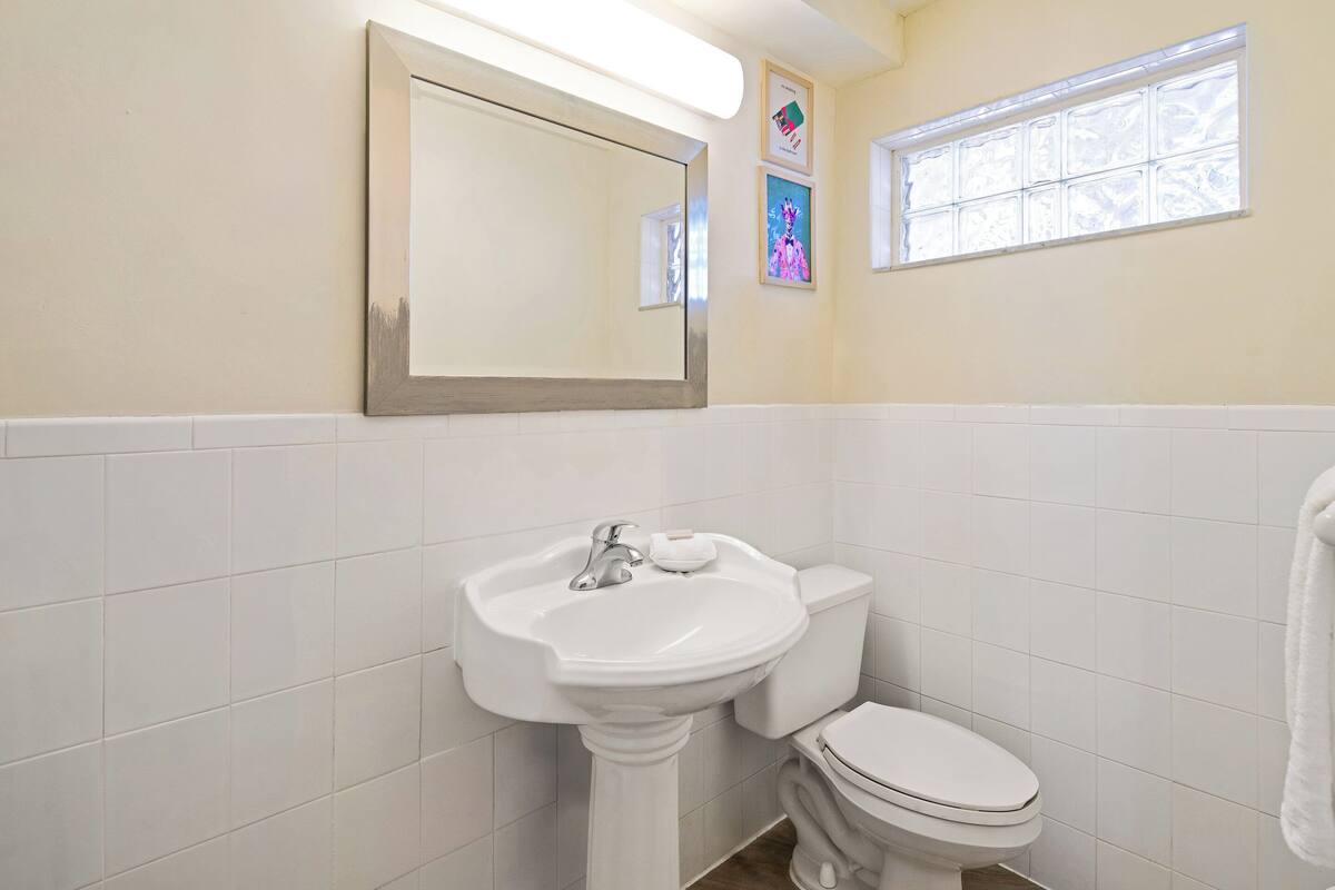 A bathroom with a pedestal sink, a toilet, a mirror, and a window.
