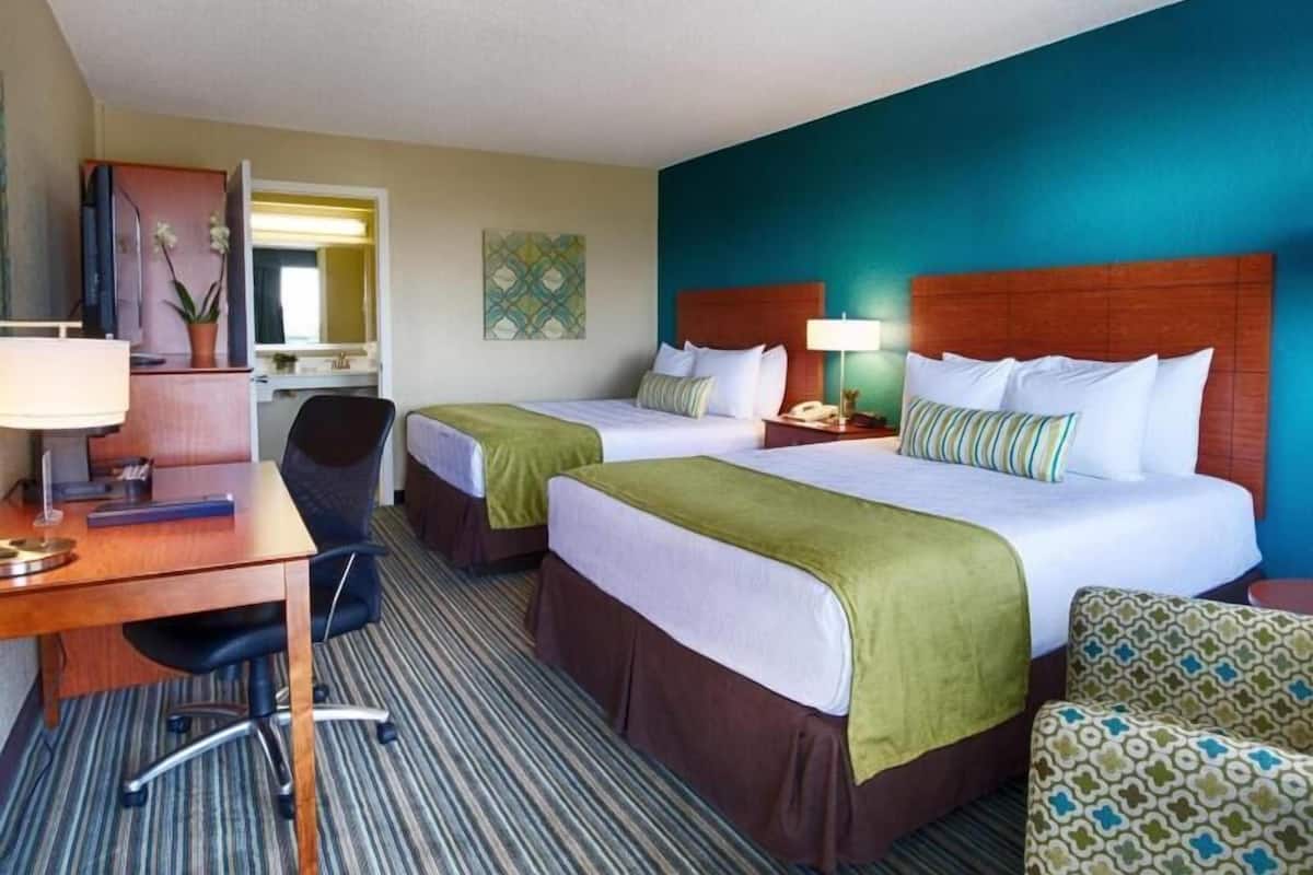 A hotel room with two beds, a desk with a chair, a lamp, and a patterned armchair.