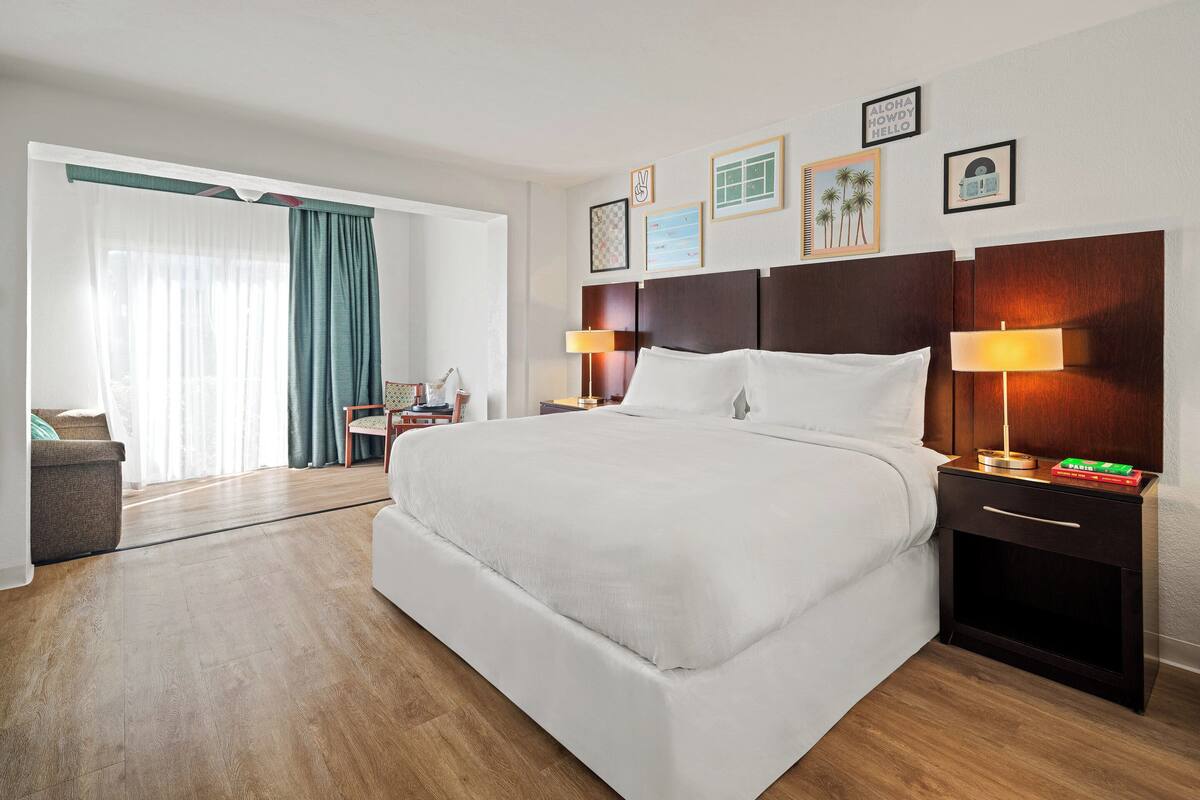 A hotel room with a large bed, two bedside tables with lamps, a wooden headboard, and a view of the outside through a window with curtains.