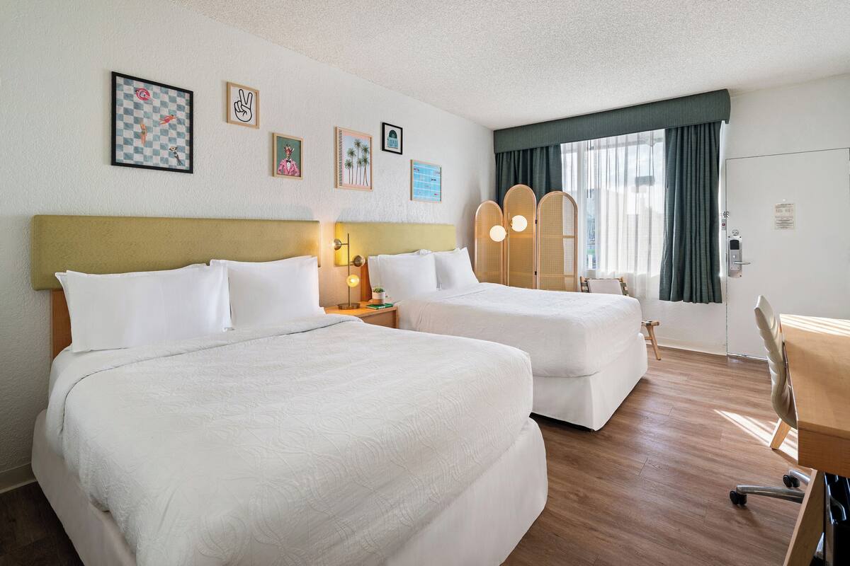 A hotel room with two beds, a desk, and a chair. There are framed pictures on the wall and a window with curtains.