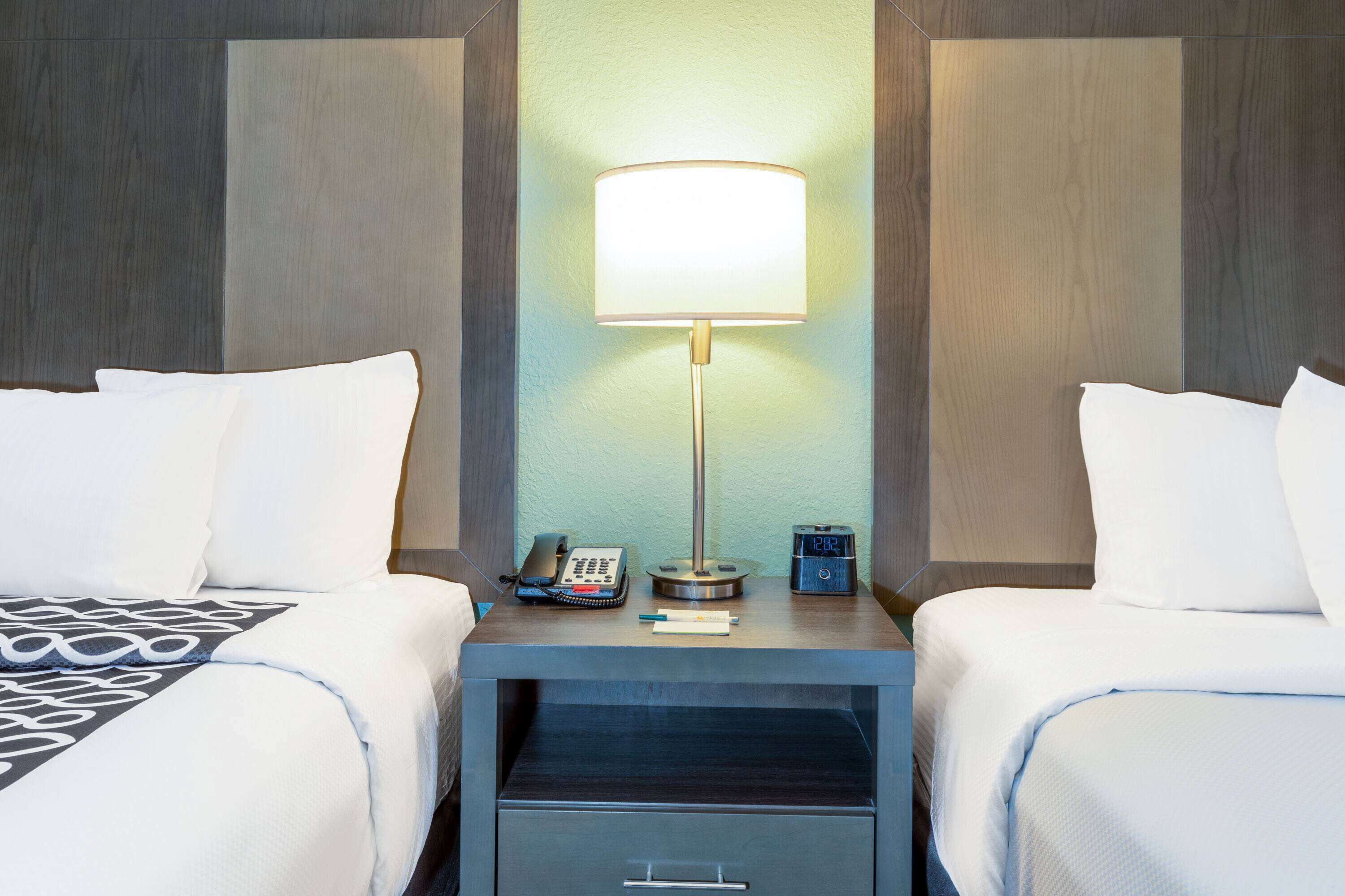 A hotel room with two beds, a nightstand with a lamp, a phone, and a digital clock.