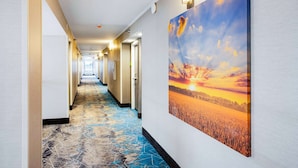 Een hotelgang met een lange gang, deuren aan beide zijden en een groot schilderij van een zonsondergang boven een veld aan de rechtermuur.