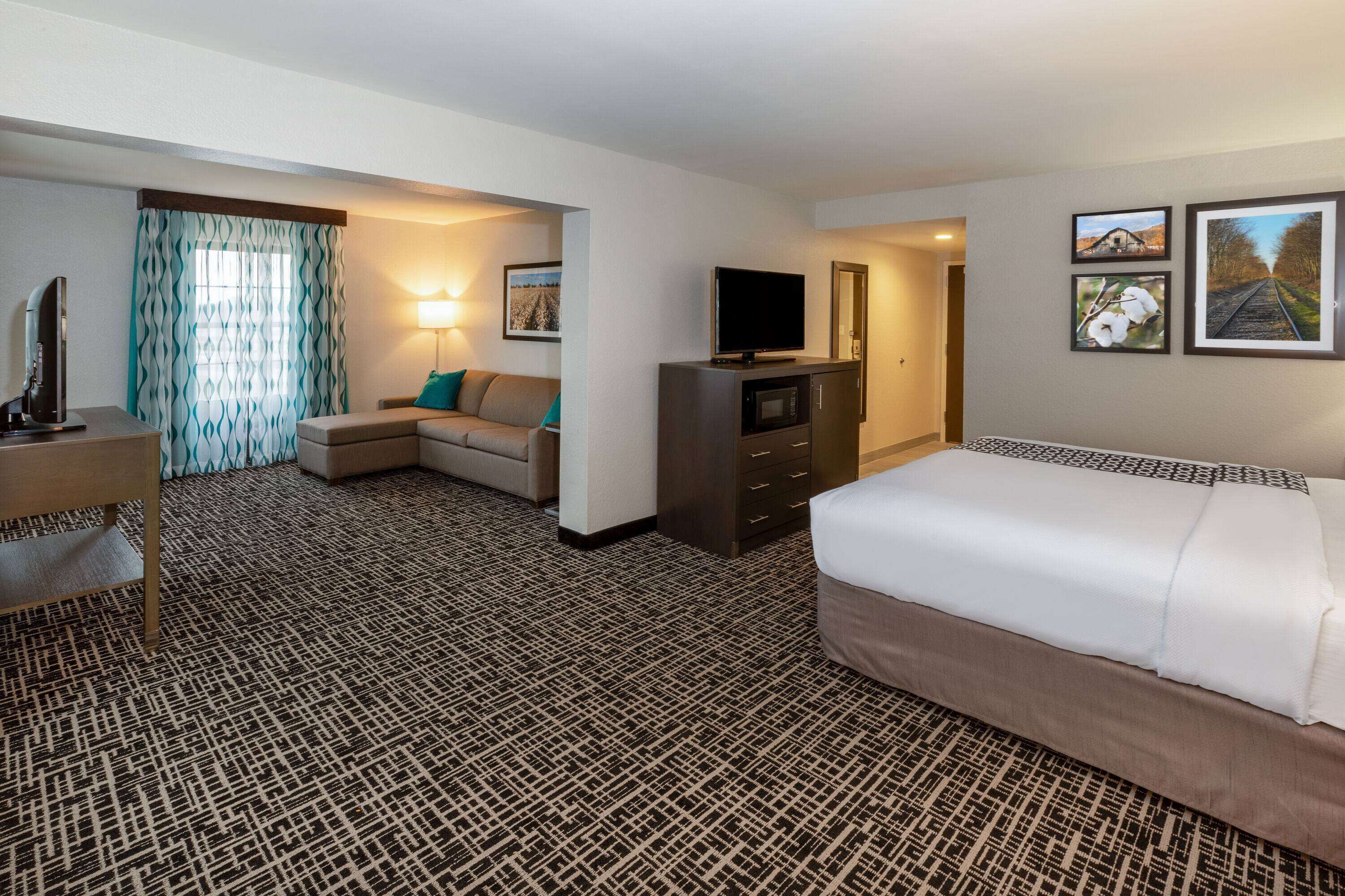A hotel room with a patterned carpet, a bed, a sofa, a TV, a desk, and a lamp.