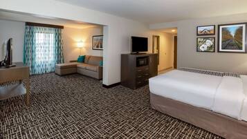 A hotel room with a patterned carpet, a bed, a sofa, a TV, a desk, and a lamp.