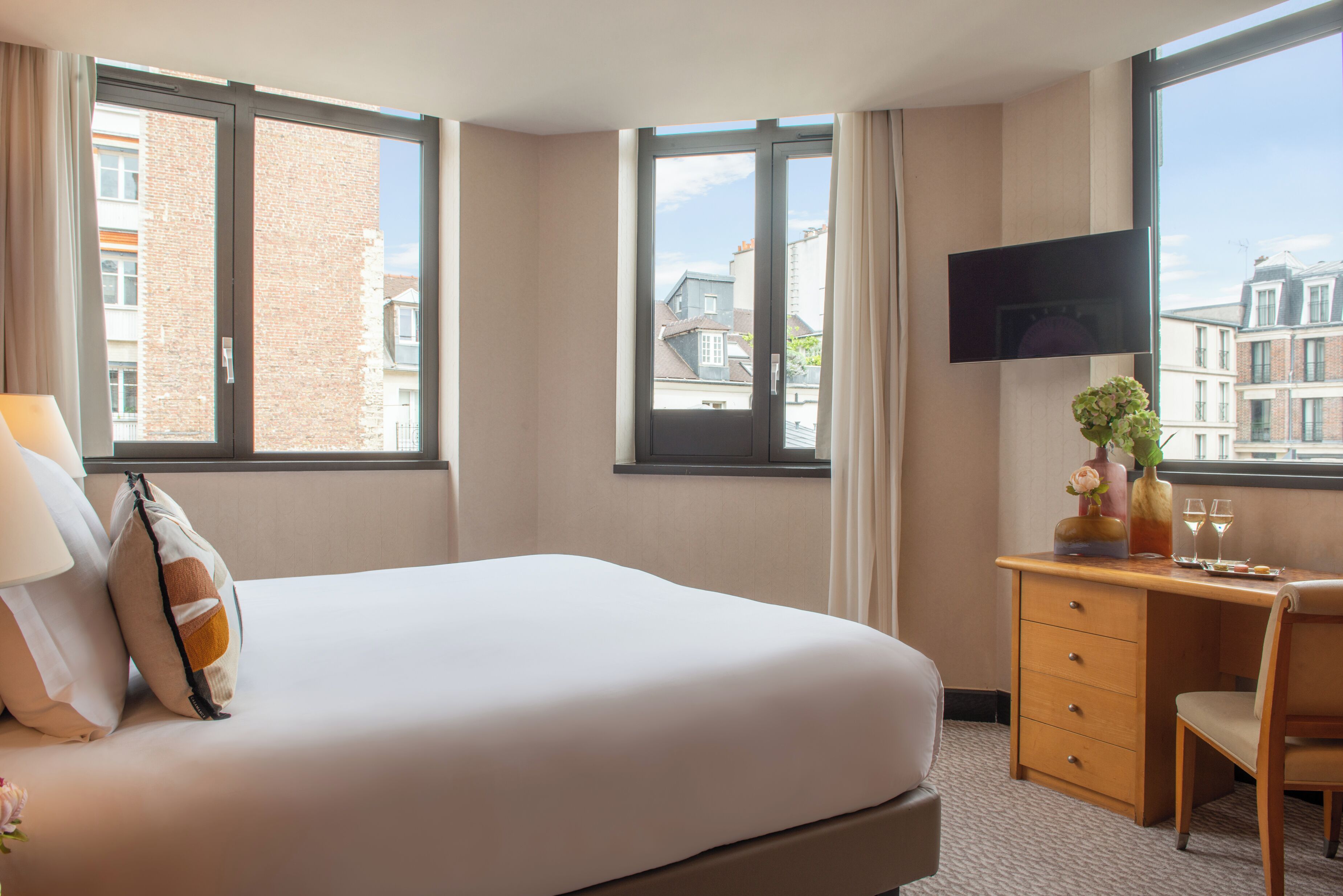 Una camera d'albergo con un letto, una scrivania, una sedia, una TV a muro e una finestra con vista sugli edifici.
