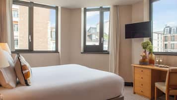 Una camera d'albergo con un letto, una scrivania, una sedia, una TV a muro e una finestra con vista sugli edifici.