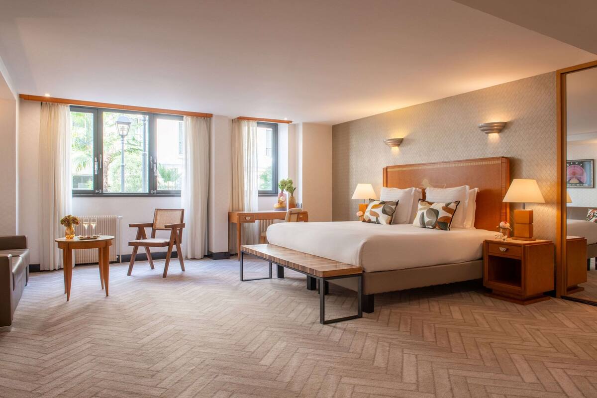 Ein Hotelzimmer mit einem großen Bett, Nachttischen, einem Schreibtisch, einem Sessel und Blick ins Freie.