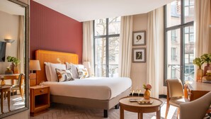 Habitación de hotel con una cama grande, un escritorio con silla, una mesa redonda con un florero y un ventanal con cortinas.