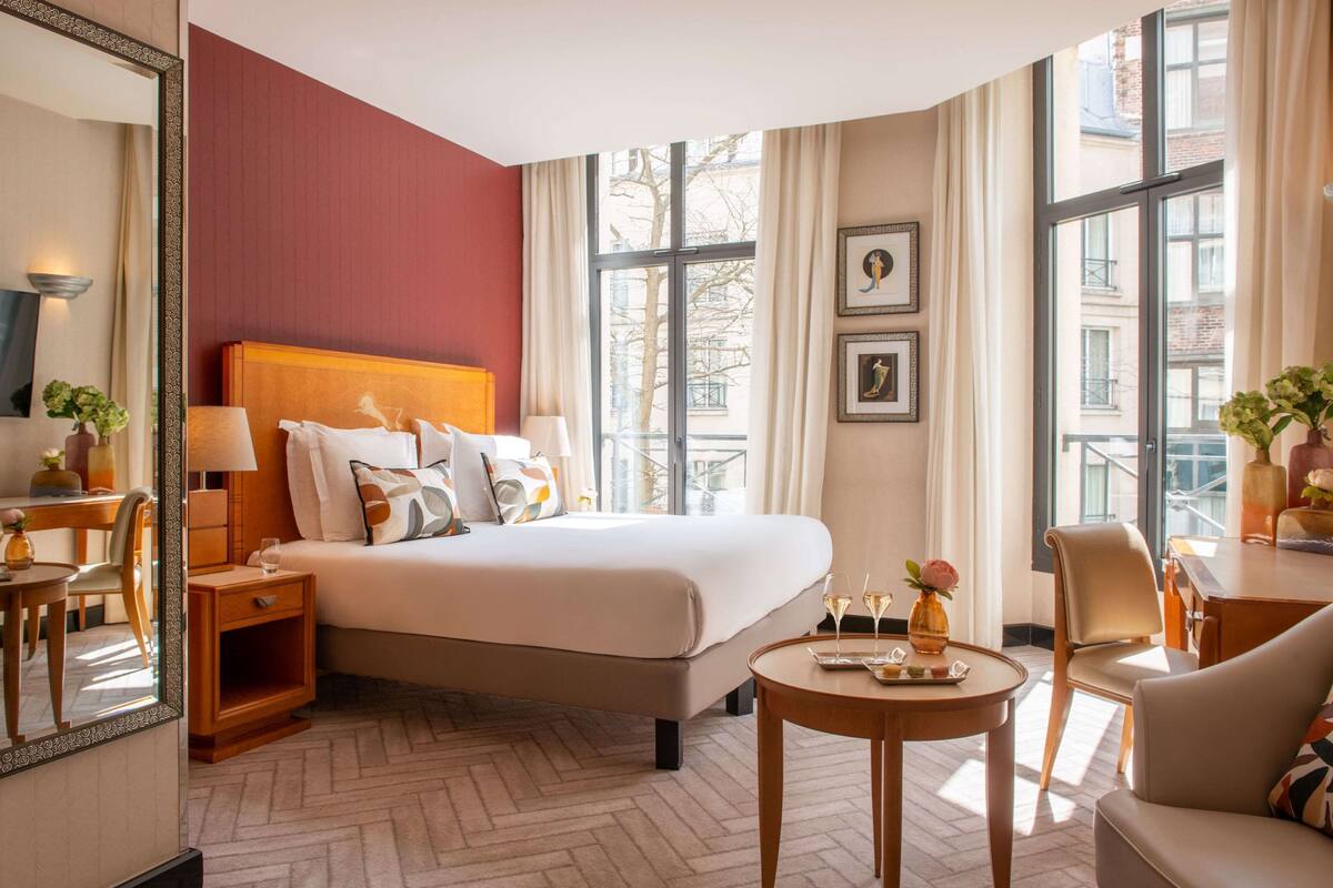Ein Hotelzimmer mit einem großen Bett, einem Schreibtisch mit Stuhl, einem runden Tisch mit Vase und einem großen Fenster mit Vorhängen.