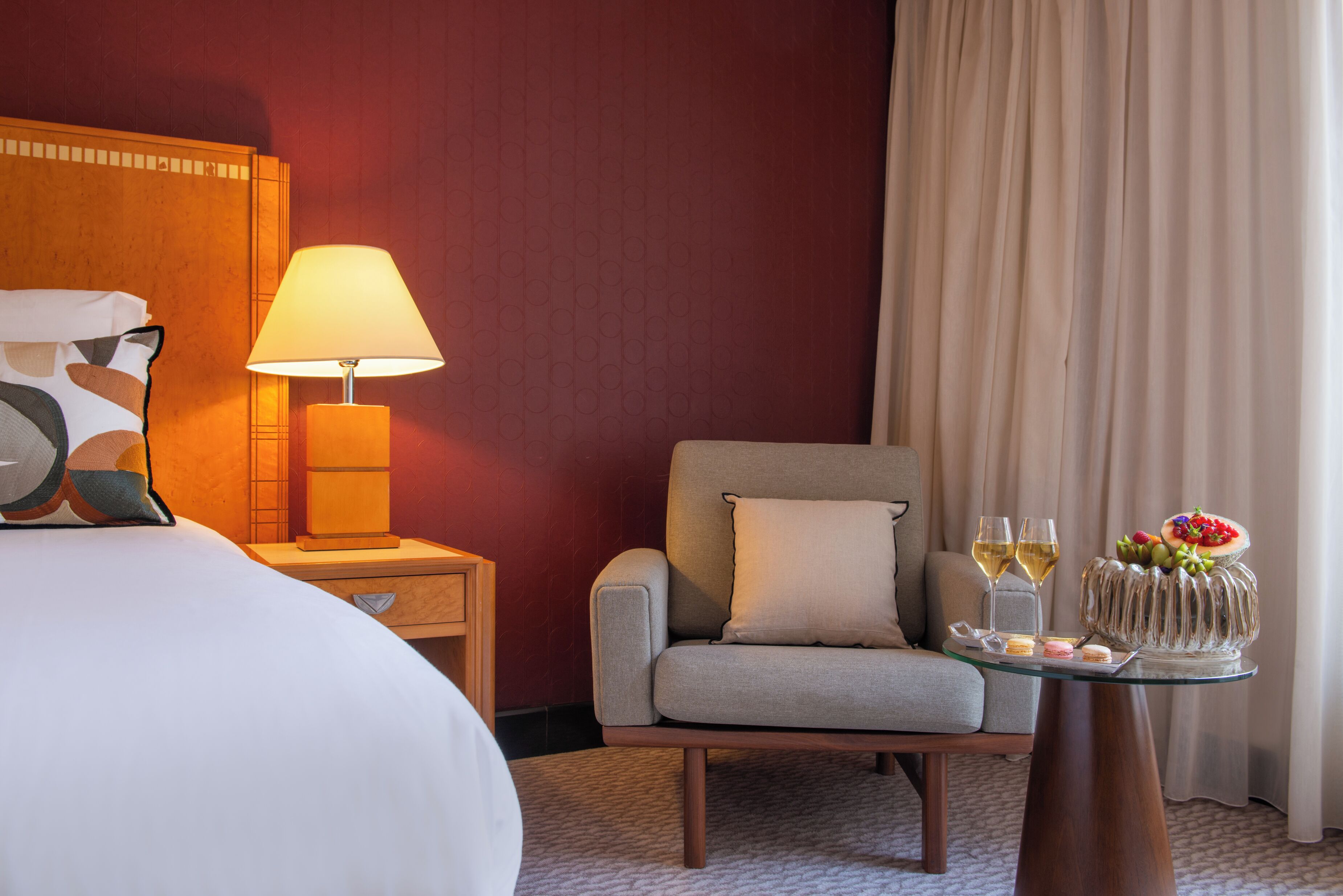 Hotelkamer met een bed, een stoel, een nachtkastje met een lamp, een glazen tafel met een schaal fruit en twee glazen wijn.