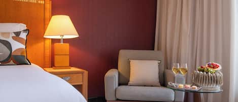 Habitación de hotel con cama, una silla, mesita de noche con lámpara, una mesa de cristal con frutero y dos copas de vino.
