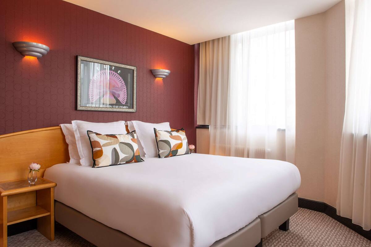 Ein Hotelzimmer mit einem großen Bett, zwei Nachttischen, einem Wandbild und einem Fenster mit durchsichtigen Vorhängen.