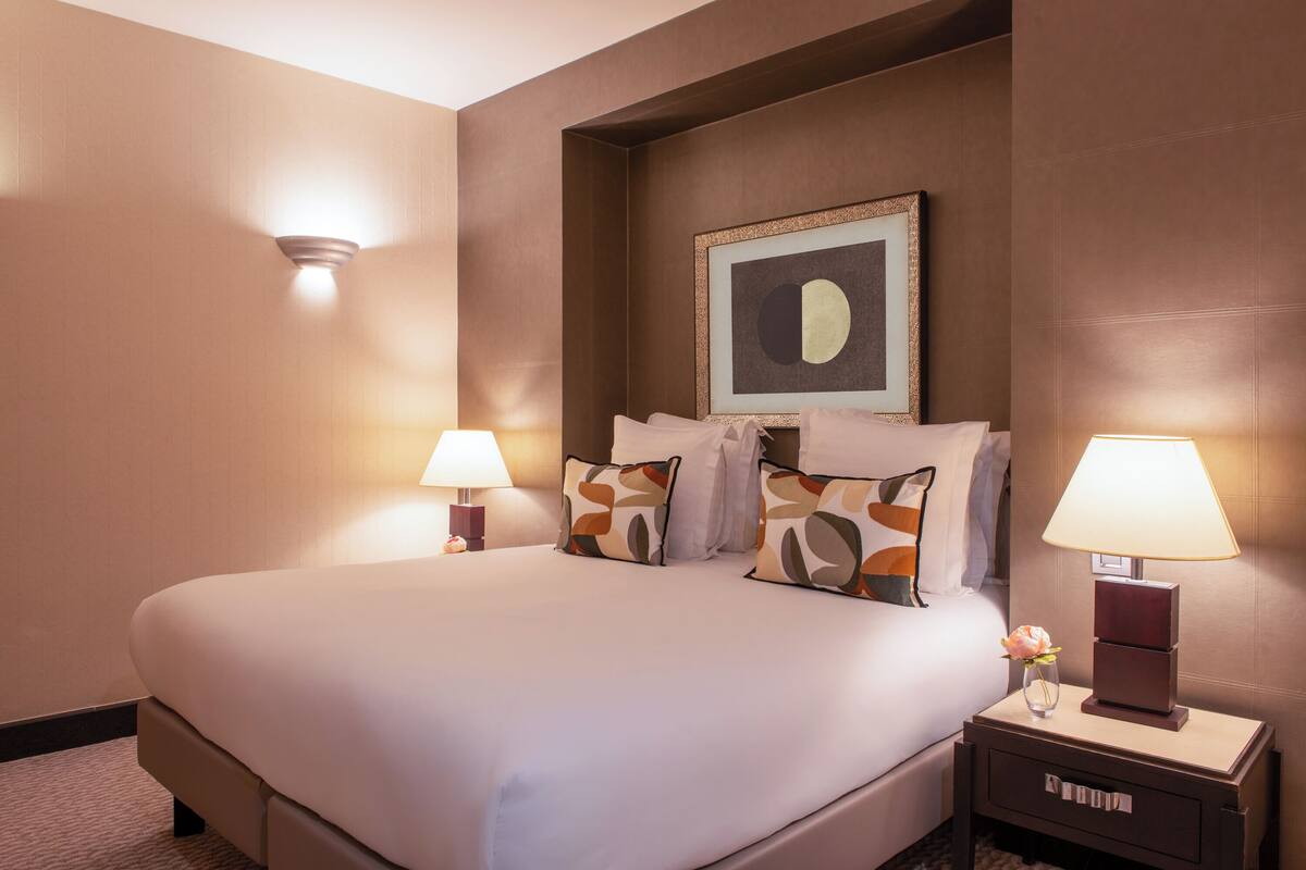Ein Hotelzimmer mit einem großen Bett, zwei Nachttischlampen, einem gerahmten Kunstwerk über dem Bett und einem Nachttisch mit einer Blumenvase.