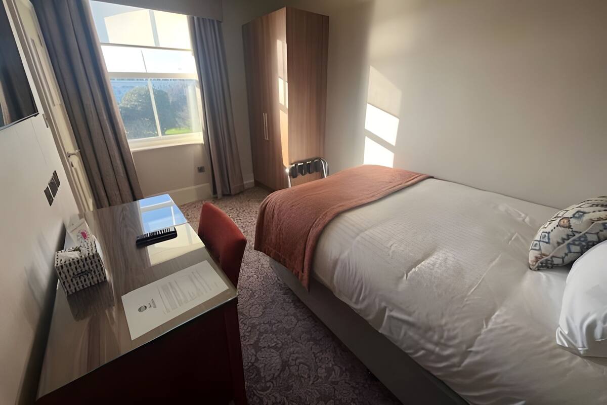 Une chambre d’hôtel avec un lit, un bureau, une chaise, une armoire et une fenêtre avec des rideaux.