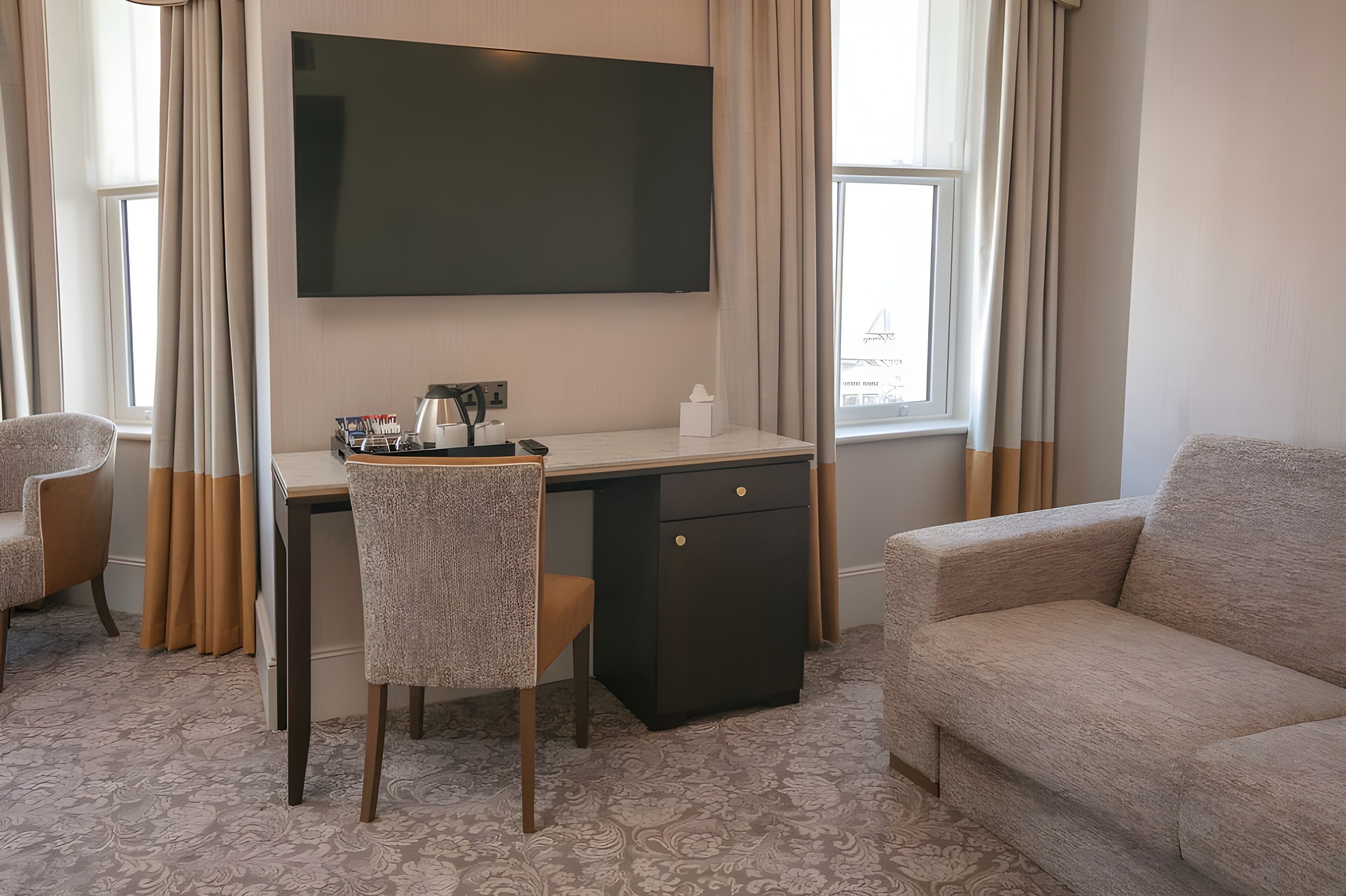 A hotel room with a flat-screen TV mounted on the wall, a desk with a chair, a small cabinet, and a sofa.
