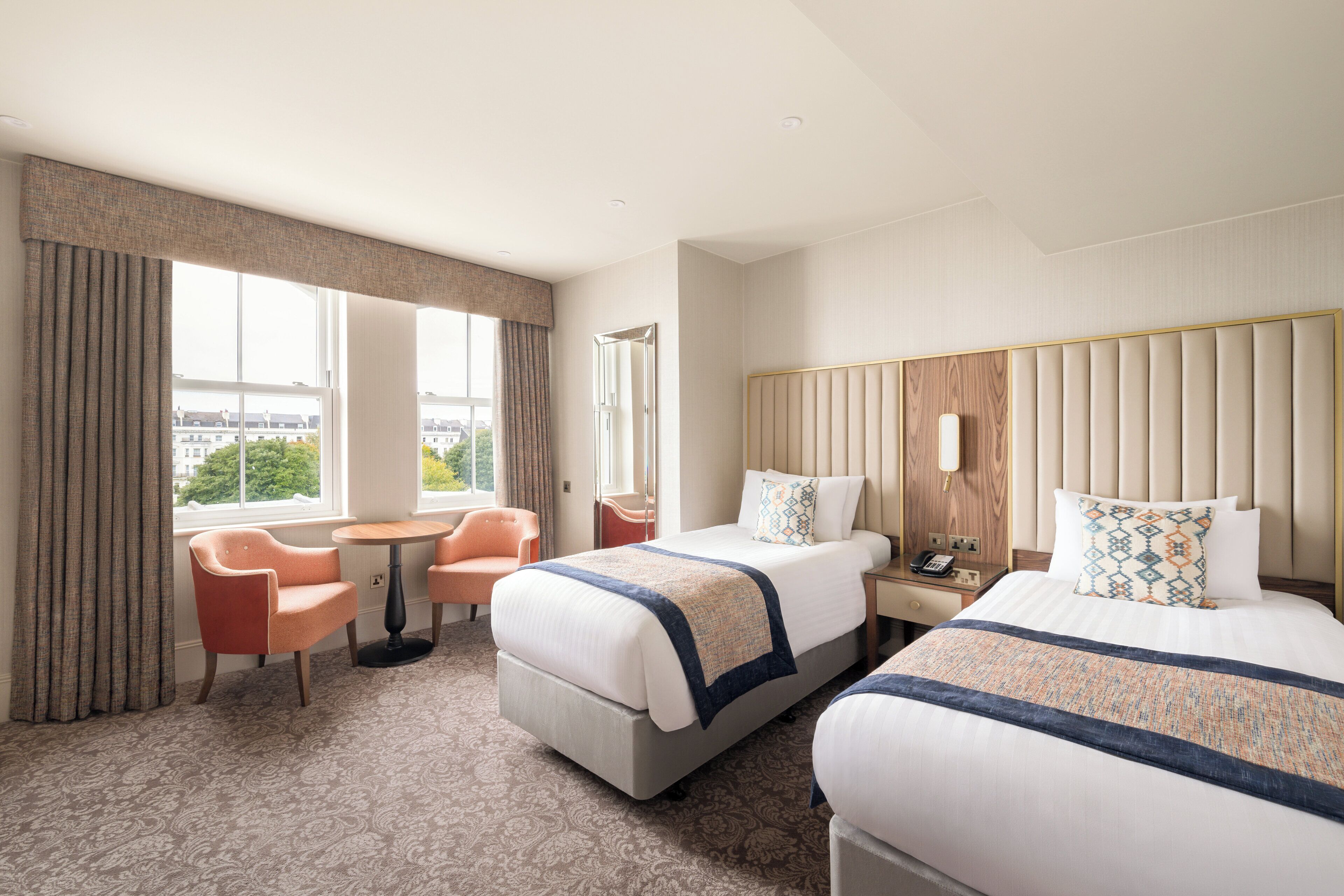 A hotel room with two beds, a small table, a chair, and a window with curtains.