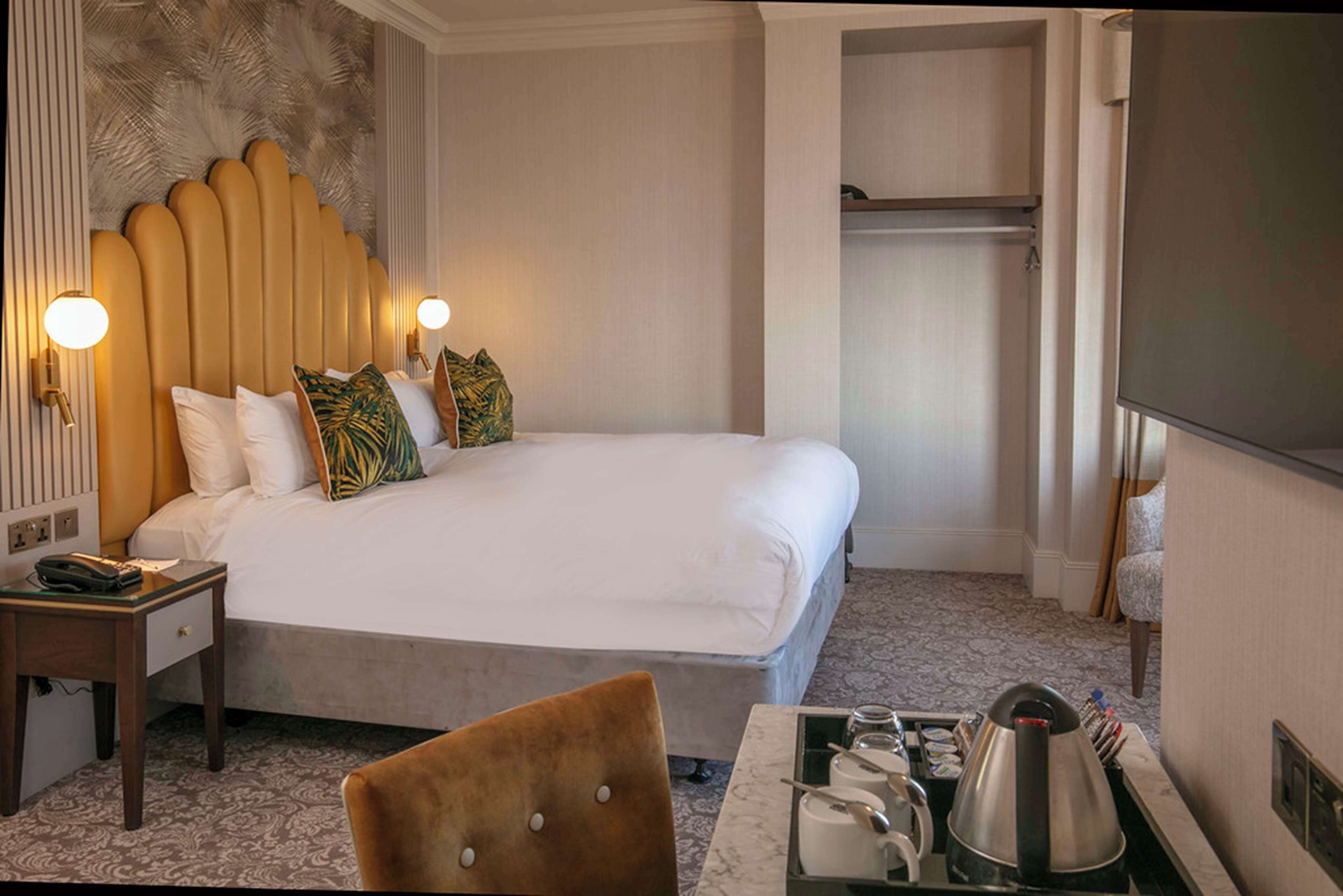 Habitación de hotel con una cama grande, una mesita de noche con lámpara, una silla, un atril con artículos de aseo y un televisor.