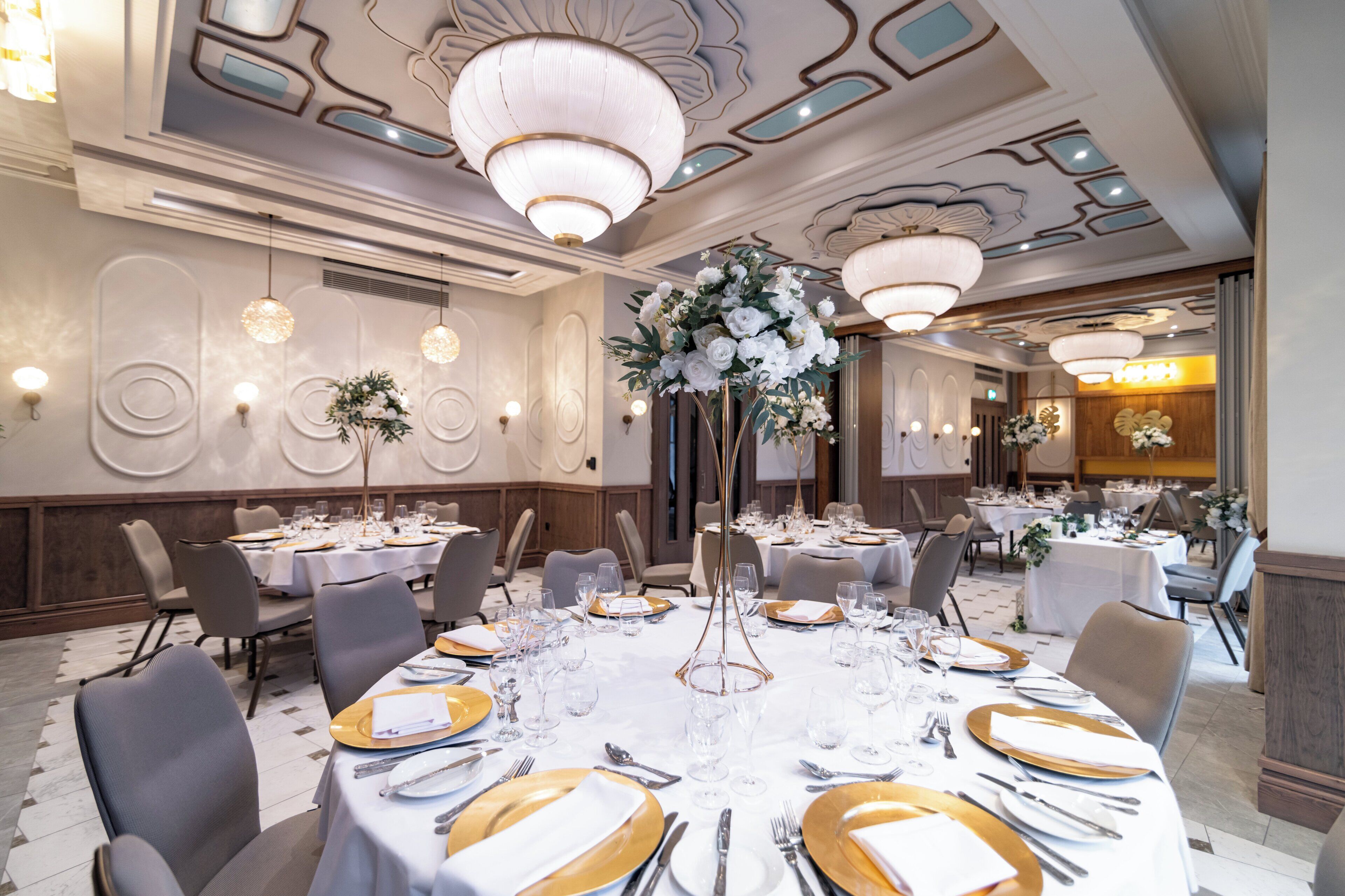 Un salón de banquetes con mesas redondas dispuestas para el servicio de comidas, con vajilla dorada y blanca, y elegantes candelabros.